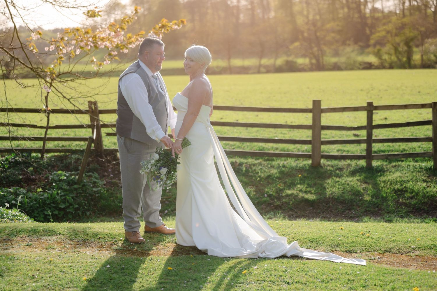 Bride and groom holding hands and gazing at each other in a field during golden hour, with warm sunlight creating a romantic glow.