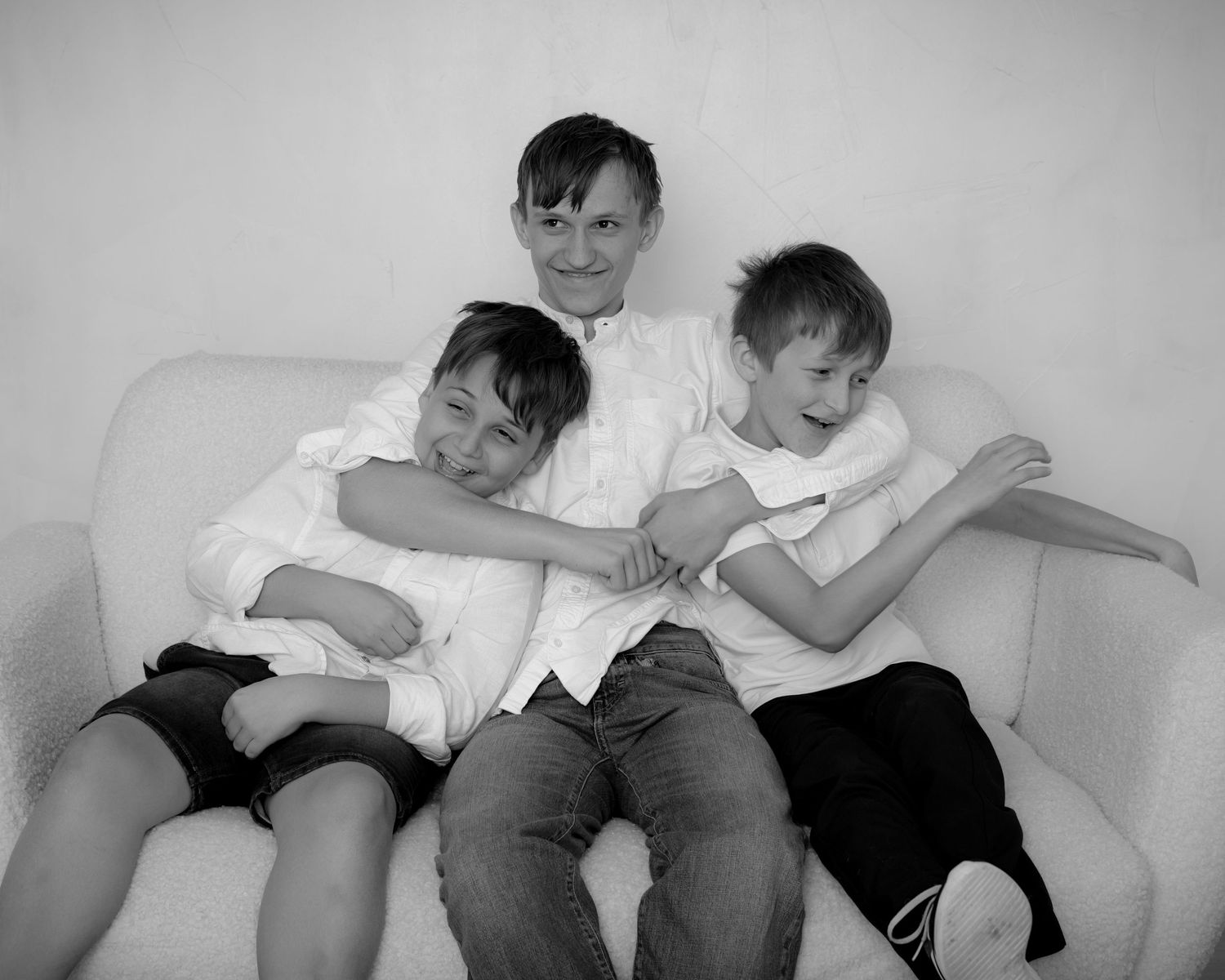Three boys in white shirts laugh and hug while sitting on a white couch.