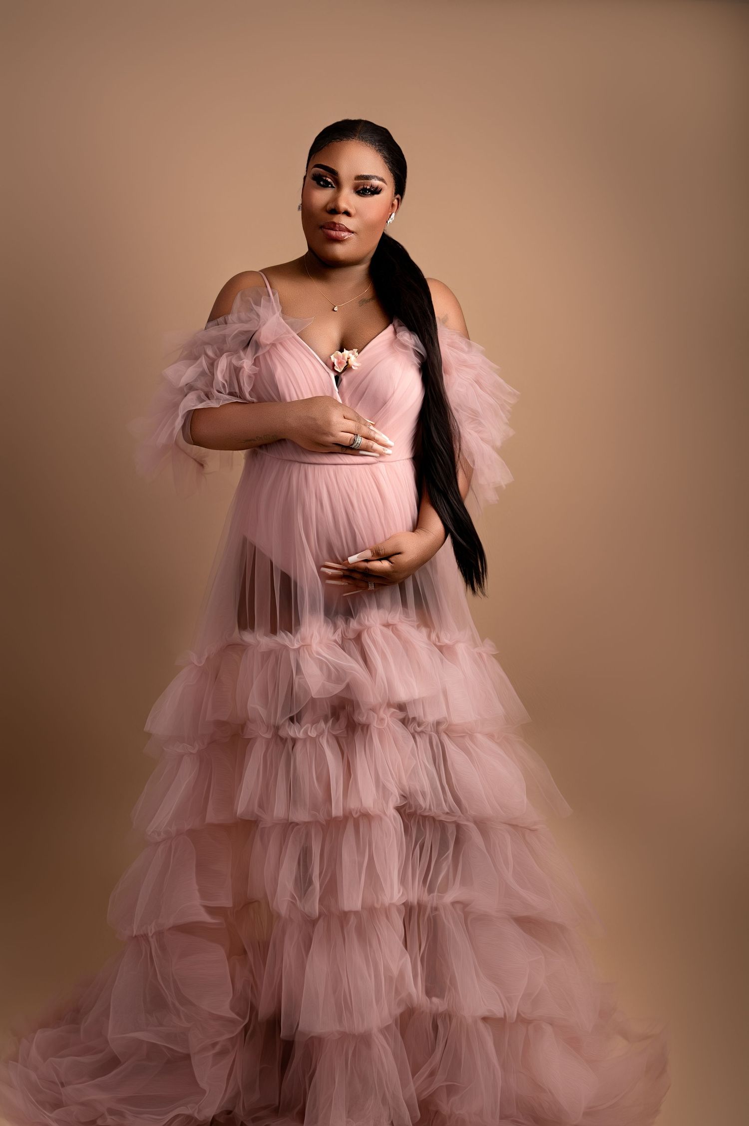 Woman facing forward in a pink layered tulle gown, standing against a neutral background, holding her belly with both hands in a serene pose