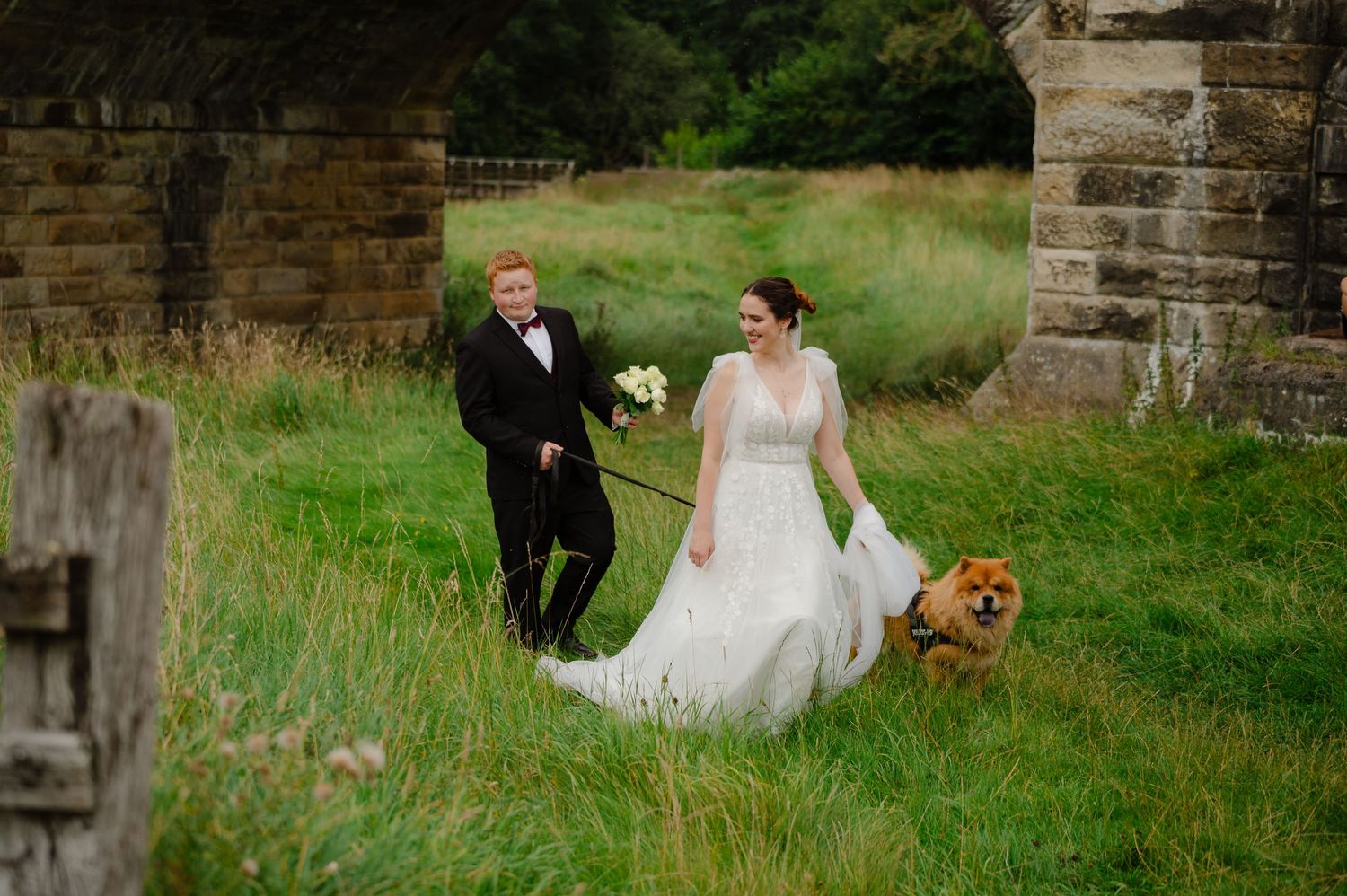 Bride and groom walking hand in hand through the grass with their dog beside them
