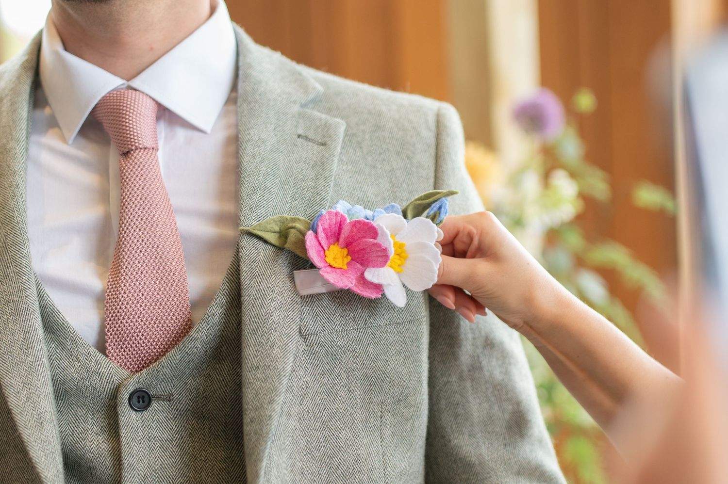 Close-up of a hand pinning a bright pink and white boutonnière onto the groom’s light grey suit jacket before the ceremony.