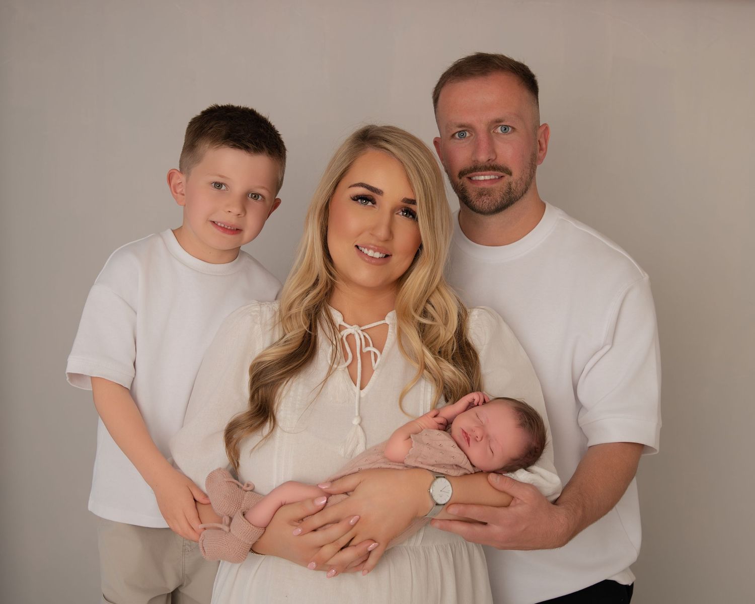 Smiling family of four posing together in white outfits, with mother holding a sleeping newborn.