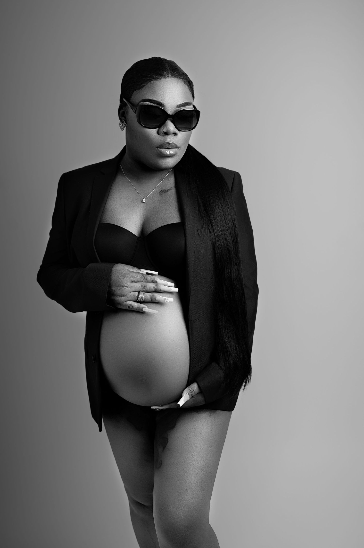 Pregnant woman in a black blazer and sunglasses, standing confidently with her bump showing against a gray background.
