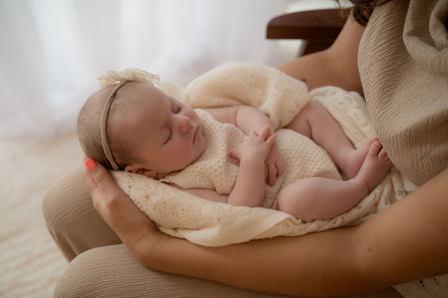 Newborn baby girl resting in mother’s arms, wrapped in a soft cream blanket with a delicate headband, in a softly lit room.