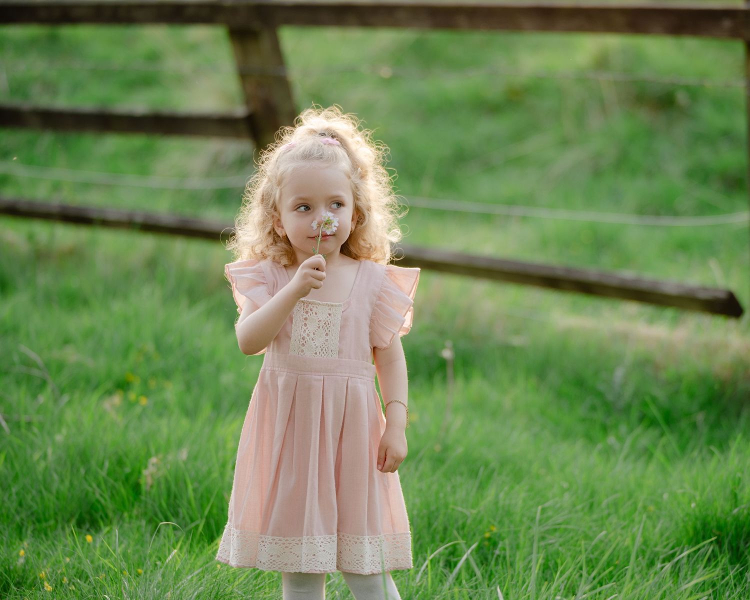 Young girl with curly blonde hair walking through tall green grass, wearing a light pink dress and holding something in her hand.