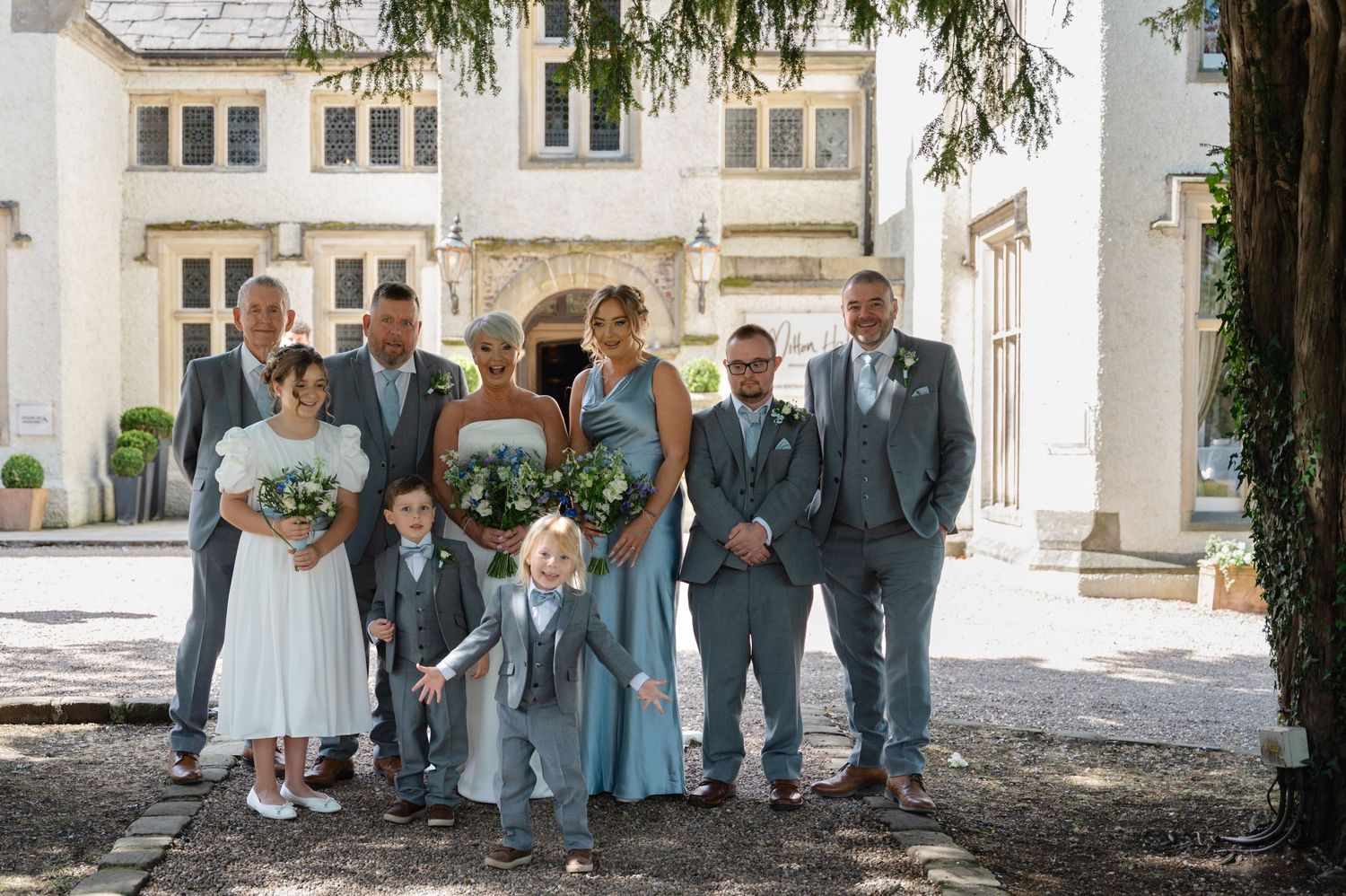 Bride and two bridesmaids seated together, smiling and raising their bouquets in celebration, in front of a large windowed venue.