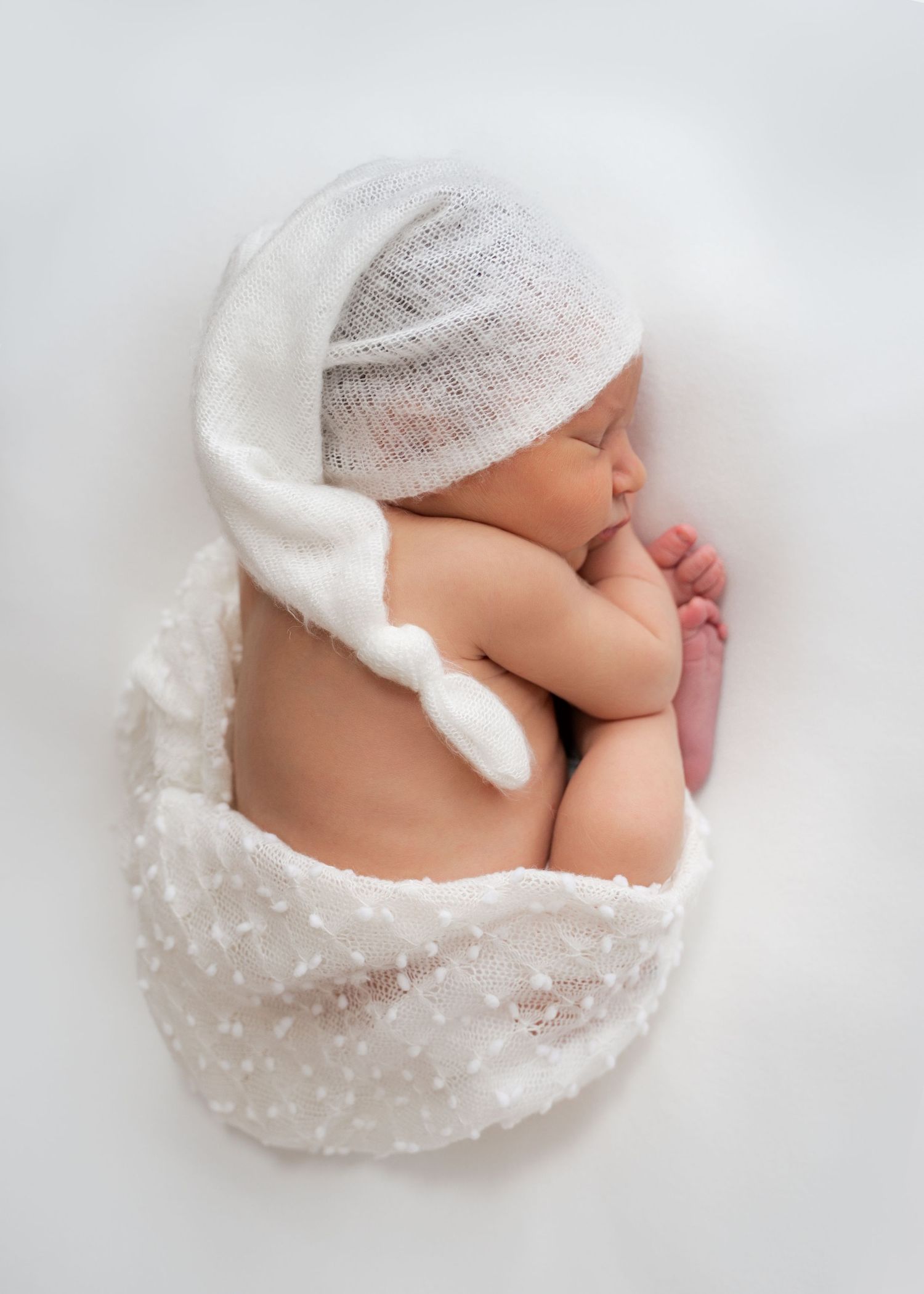 Newborn wrapped in white fabric sleeping on a white backdrop.”