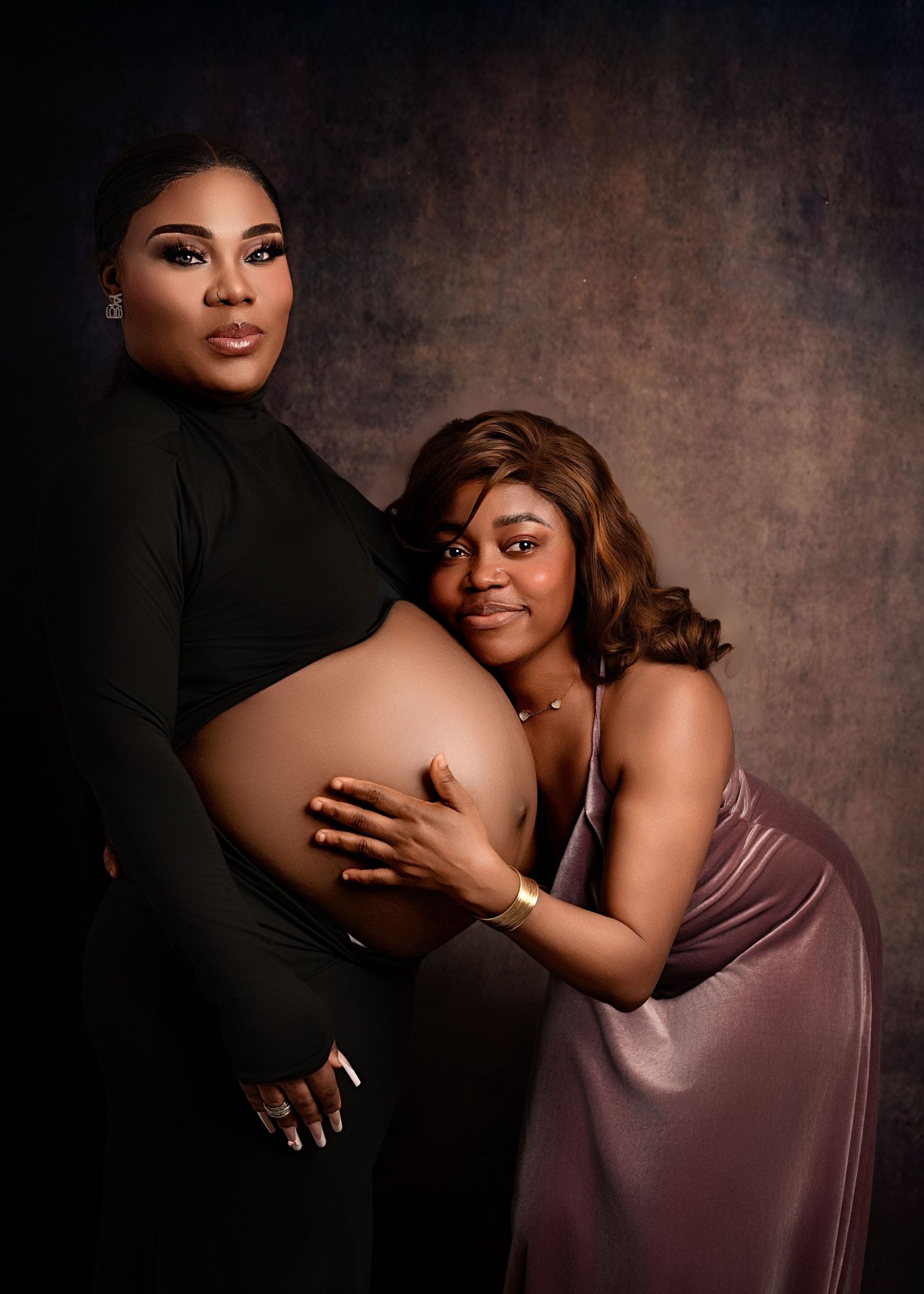 Maternity portrait of a woman in black holding her belly, sitting beside a young girl in a lavender dress who is resting her head on