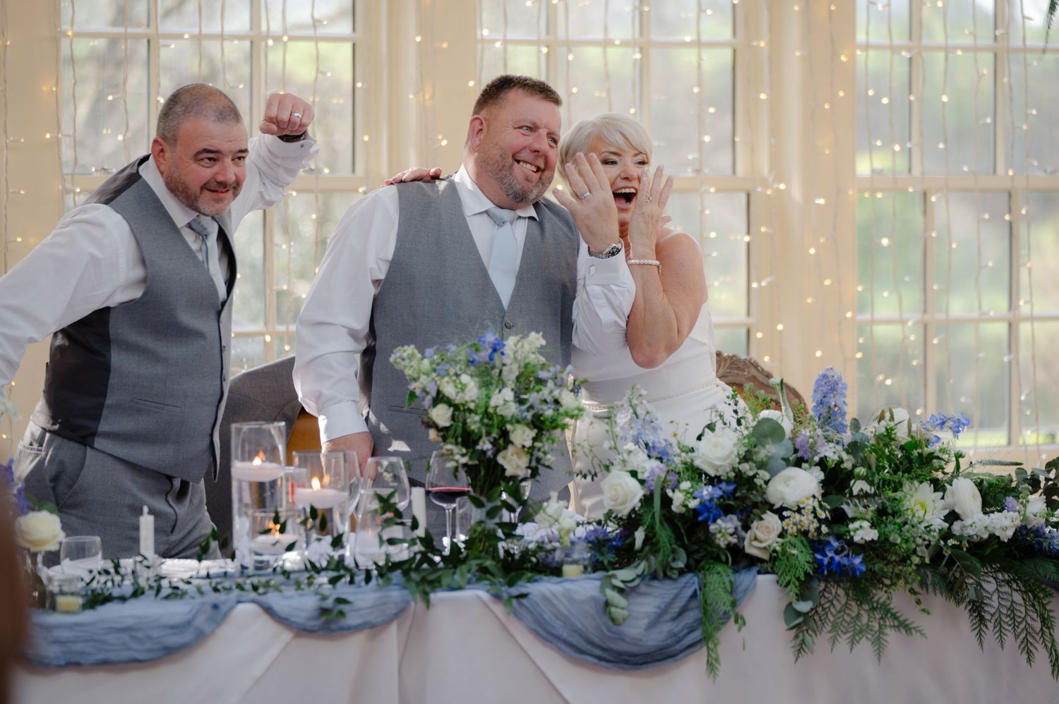 Wedding party cheering and laughing at the head table during the reception, decorated with white and blue florals and tulle draping.