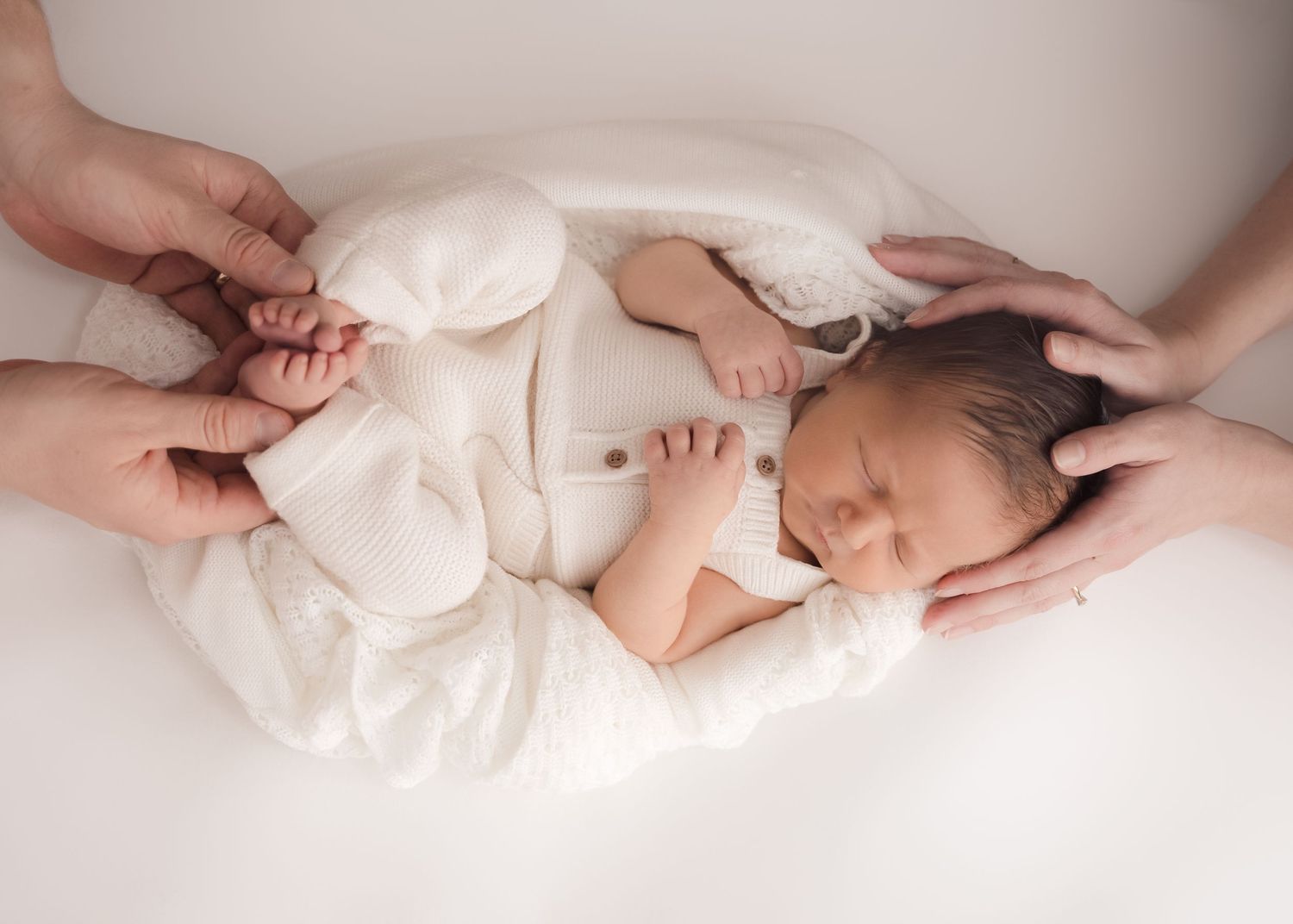Newborn wrapped in white surrounded by parents’ hands.