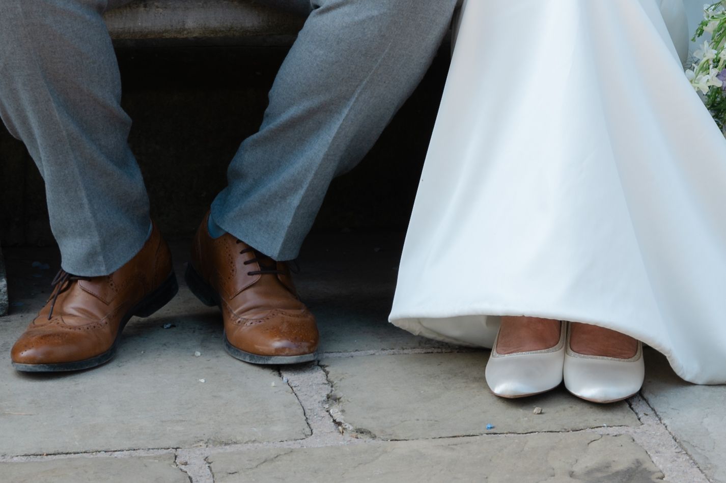 Close-up of bride and groom’s feet sitting side by side on a stone step, showing the bride’s white heels and dress hem beside the groom’s