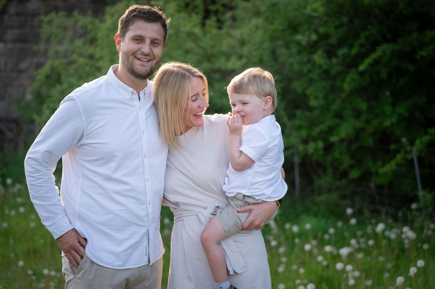 Family of three standing together in a sunlit field, all dressed in white, smiling warmly with a background of green trees and dandelions.