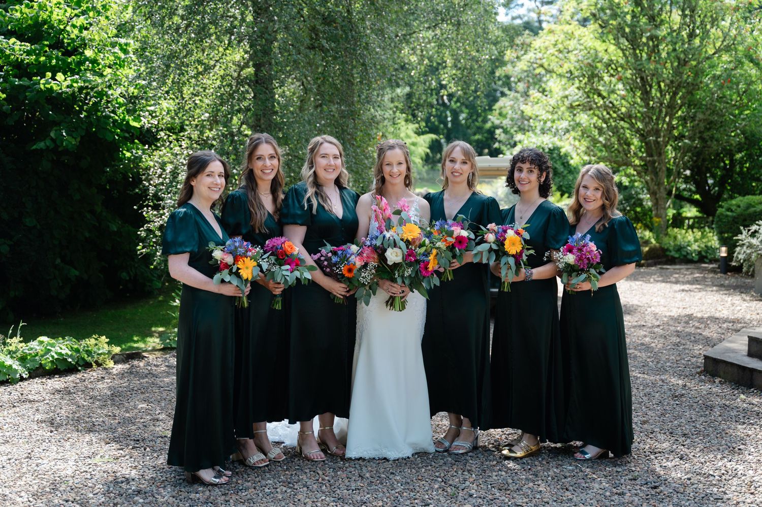 Bride with six bridesmaids in black dresses, holding colourful bouquets in a garden setting