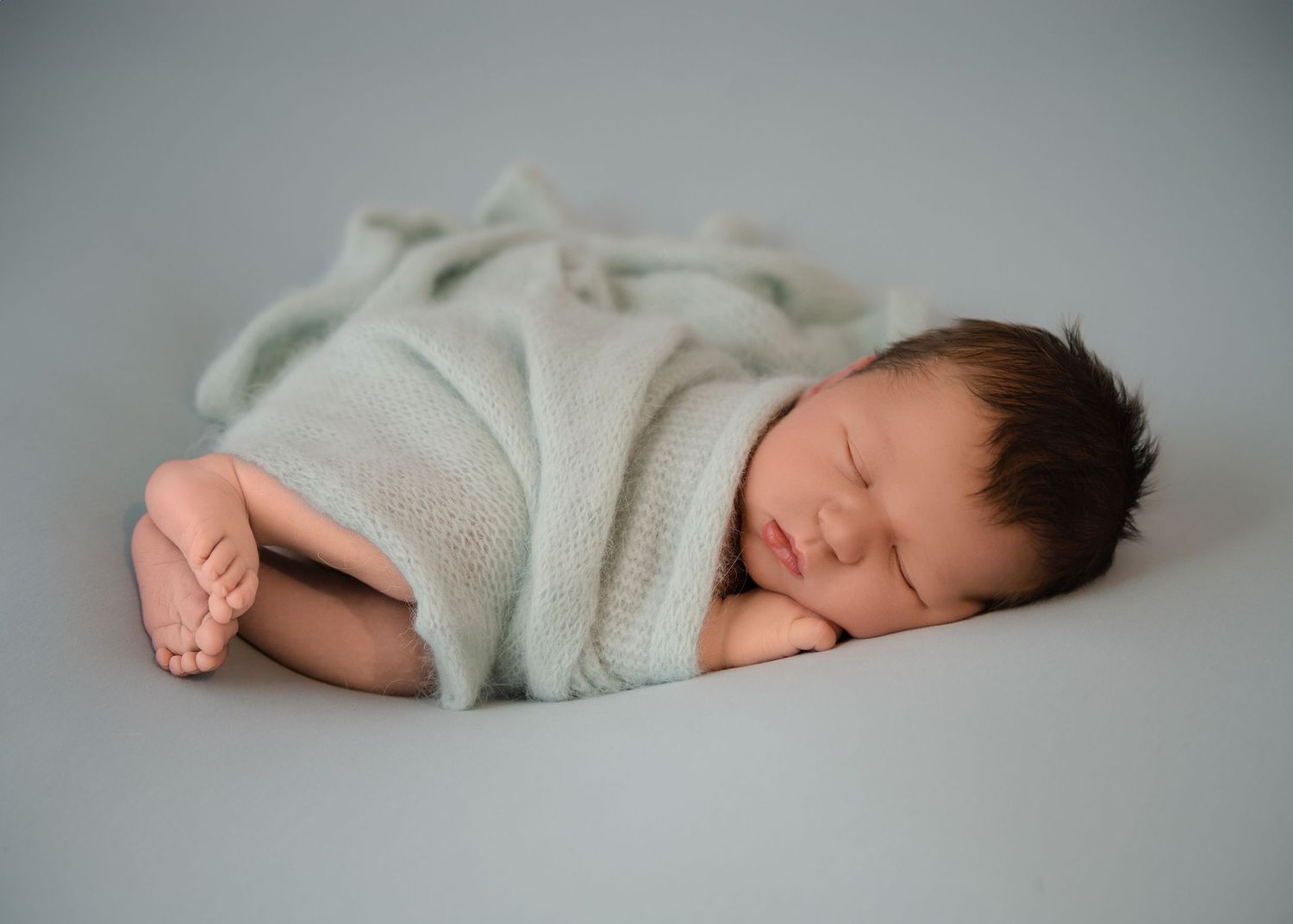 Newborn baby resting on a grey background, snuggled in a soft grey blanket with eyes closed.