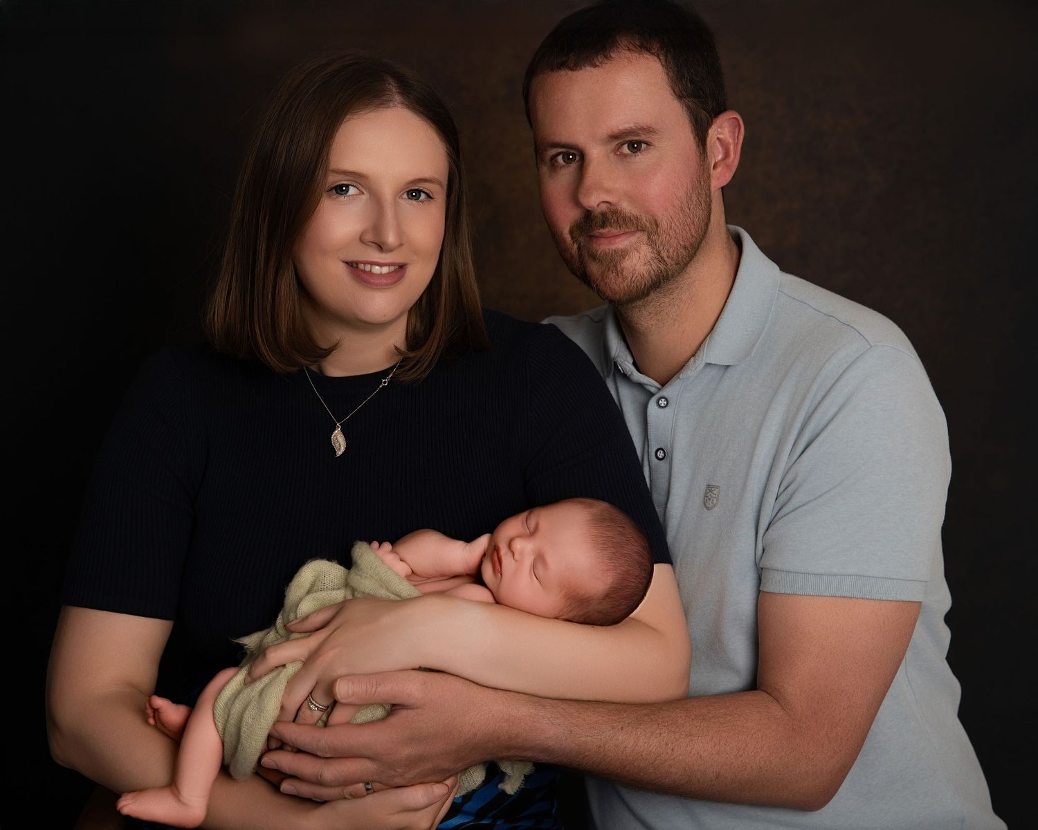 Mother and father cradling their newborn together, seated and smiling warmly at the camera.