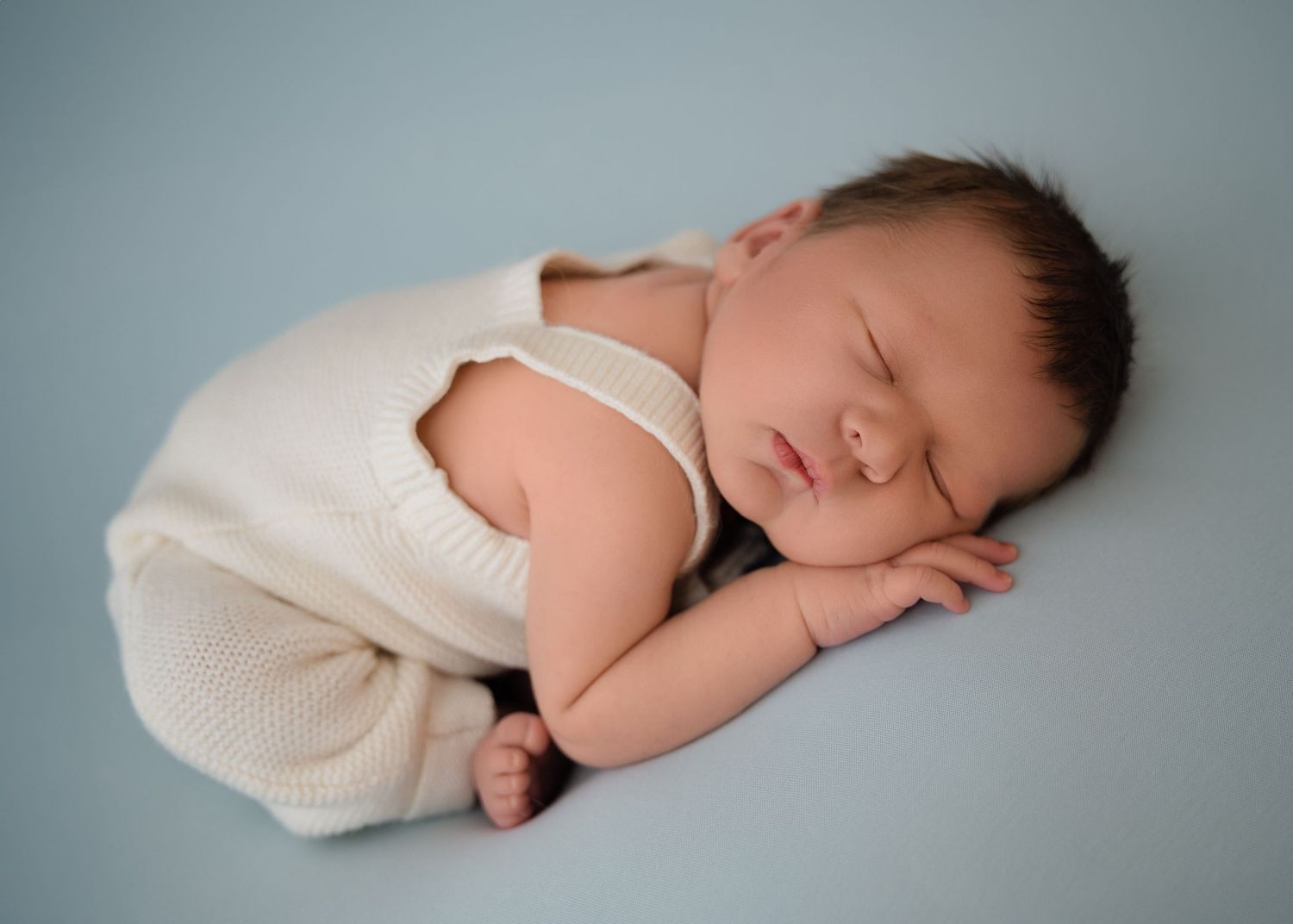 Newborn baby dressed in a cream outfit, sleeping in a curled-up position on a soft blue surface.