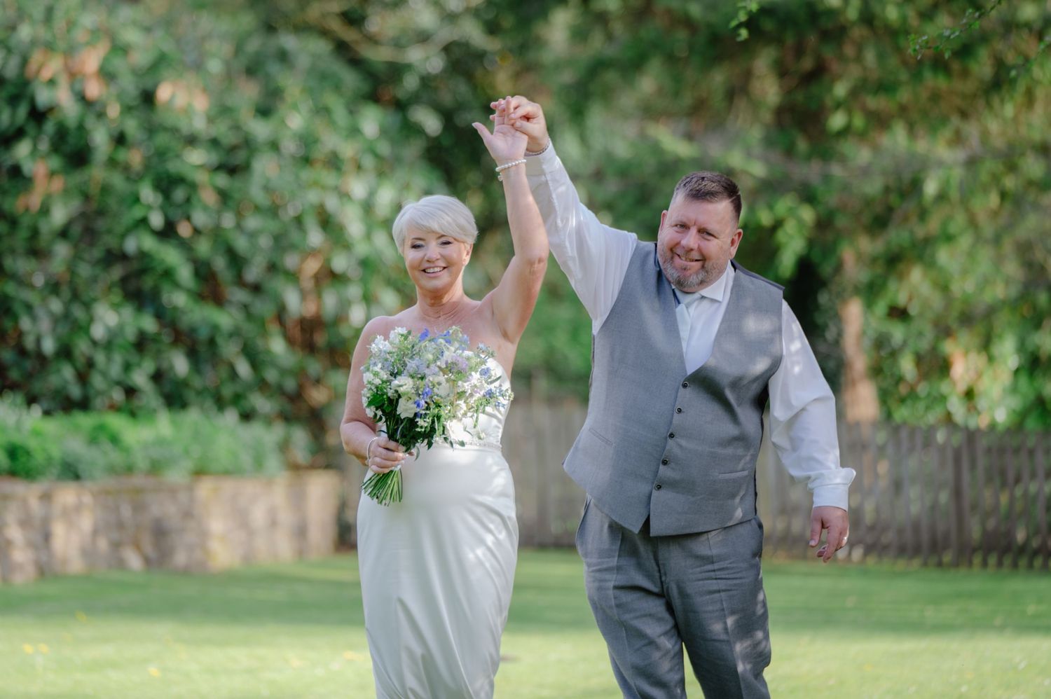 Bride and groom walking across a grassy lawn, joyfully raising their joined hands in celebration after the ceremony.