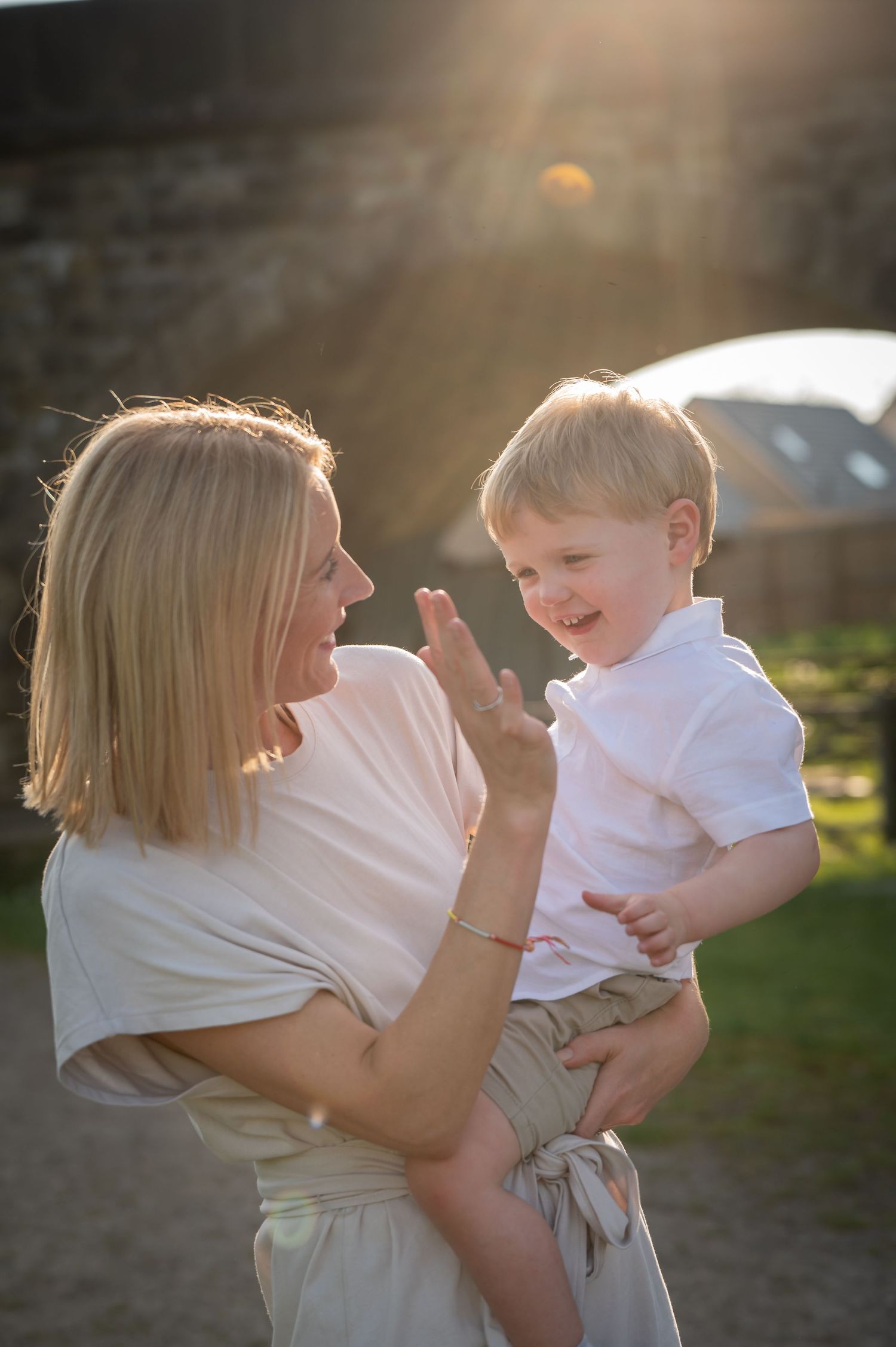 Mother holding her toddler in soft golden hour light, both dressed in light clothing, sharing a candid moment of joy.