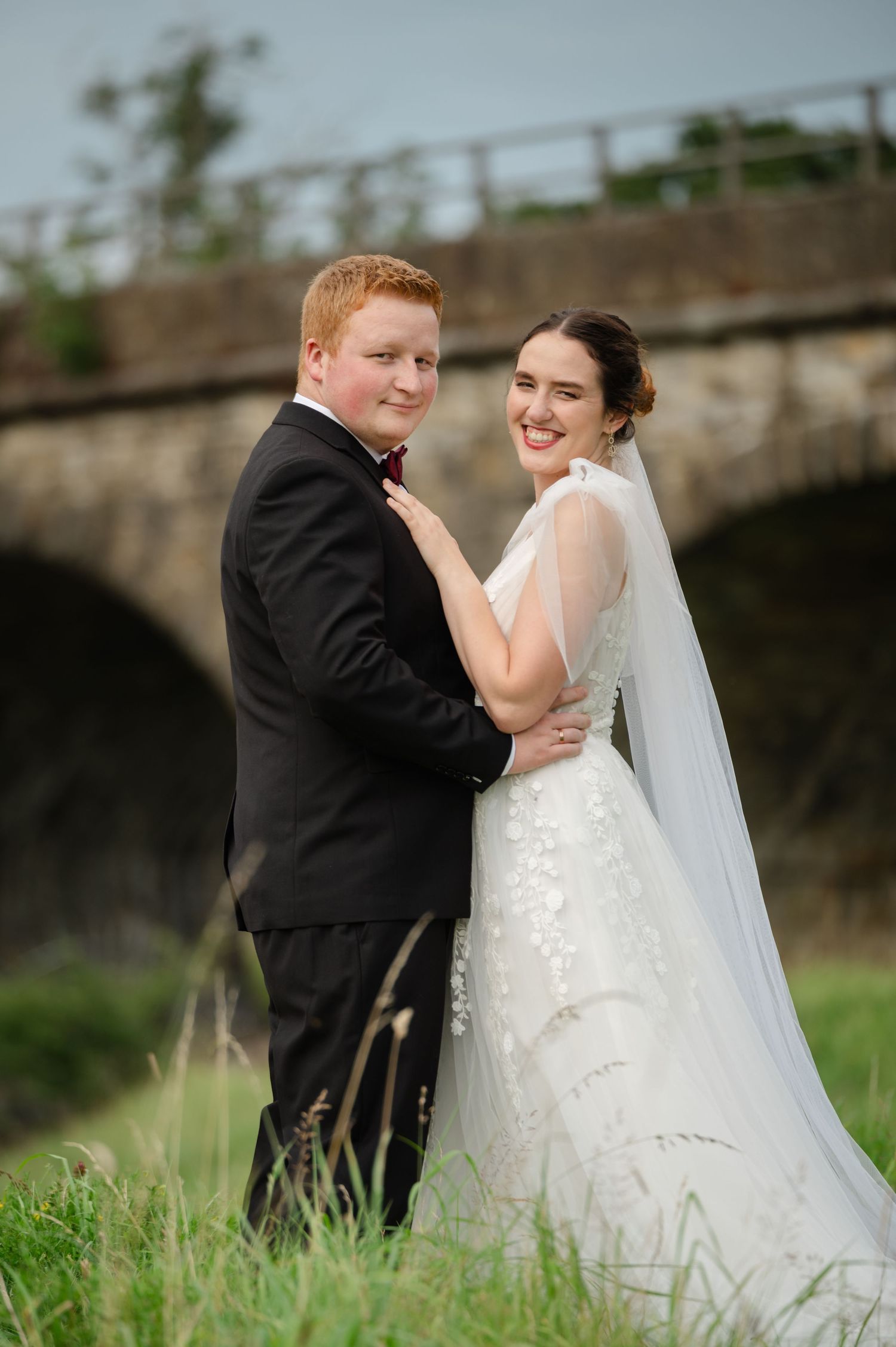 Bride and groom standing in tall grass in front of a stone viaduct