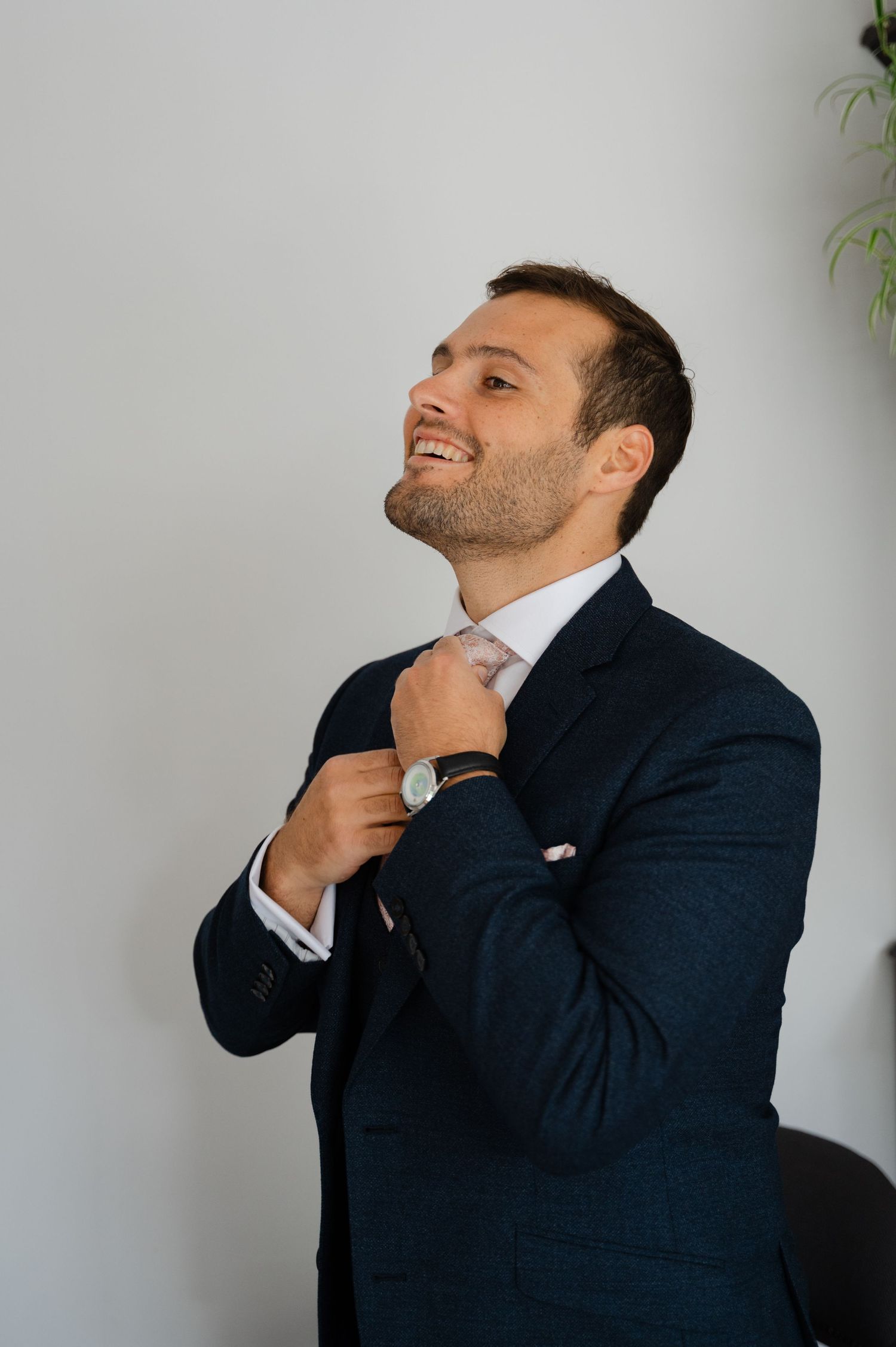 Groom smiling while adjusting his bowtie in front of a plain white wall