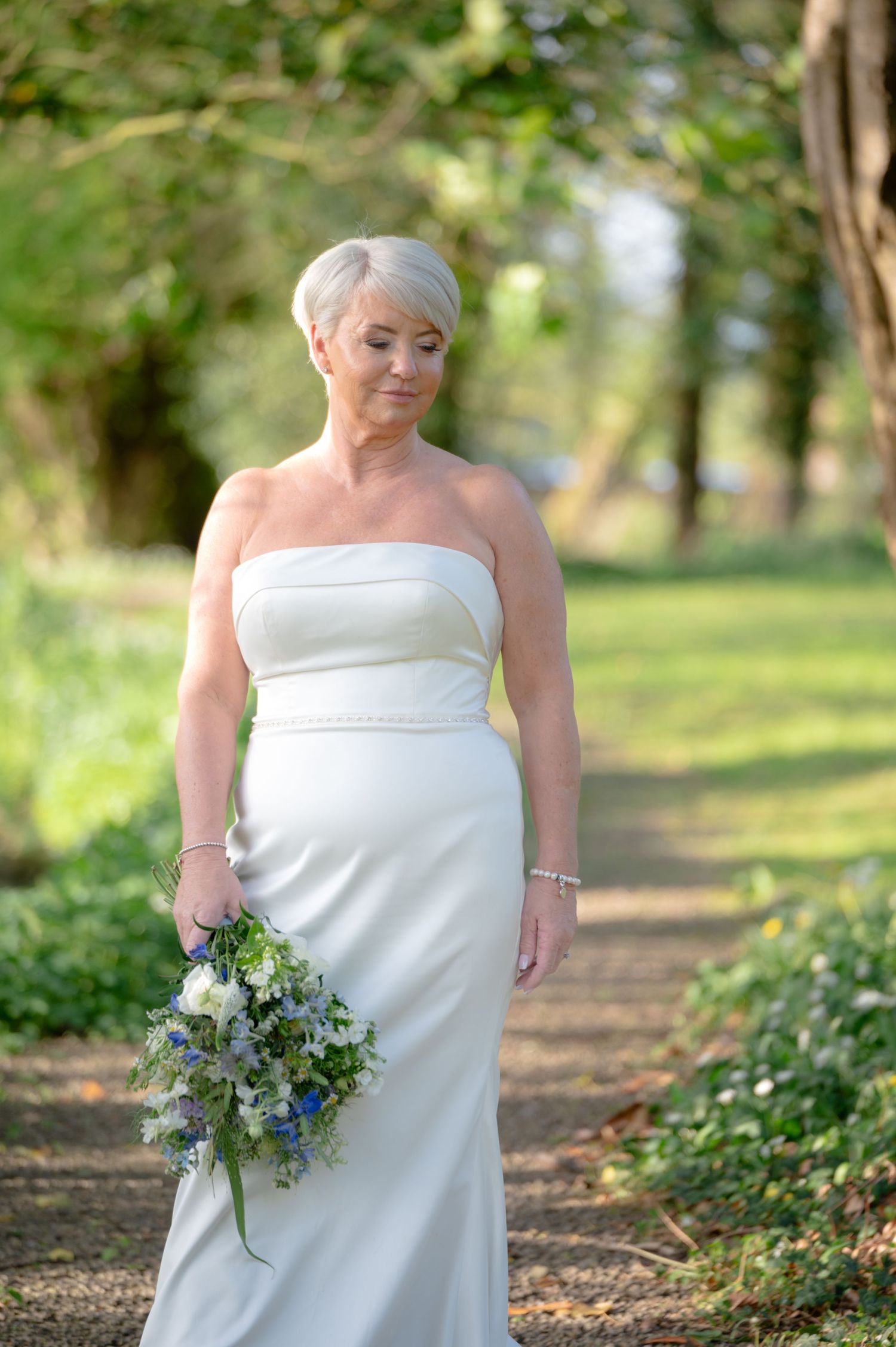 Bride standing in a sunlit woodland path, holding a bouquet and smiling peacefully in a strapless white wedding gown.