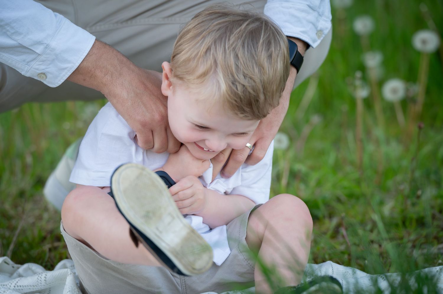 Young boy laughing joyfully while being tickled by a parent in a grassy field, sitting on a blanket surrounded by dandelions.