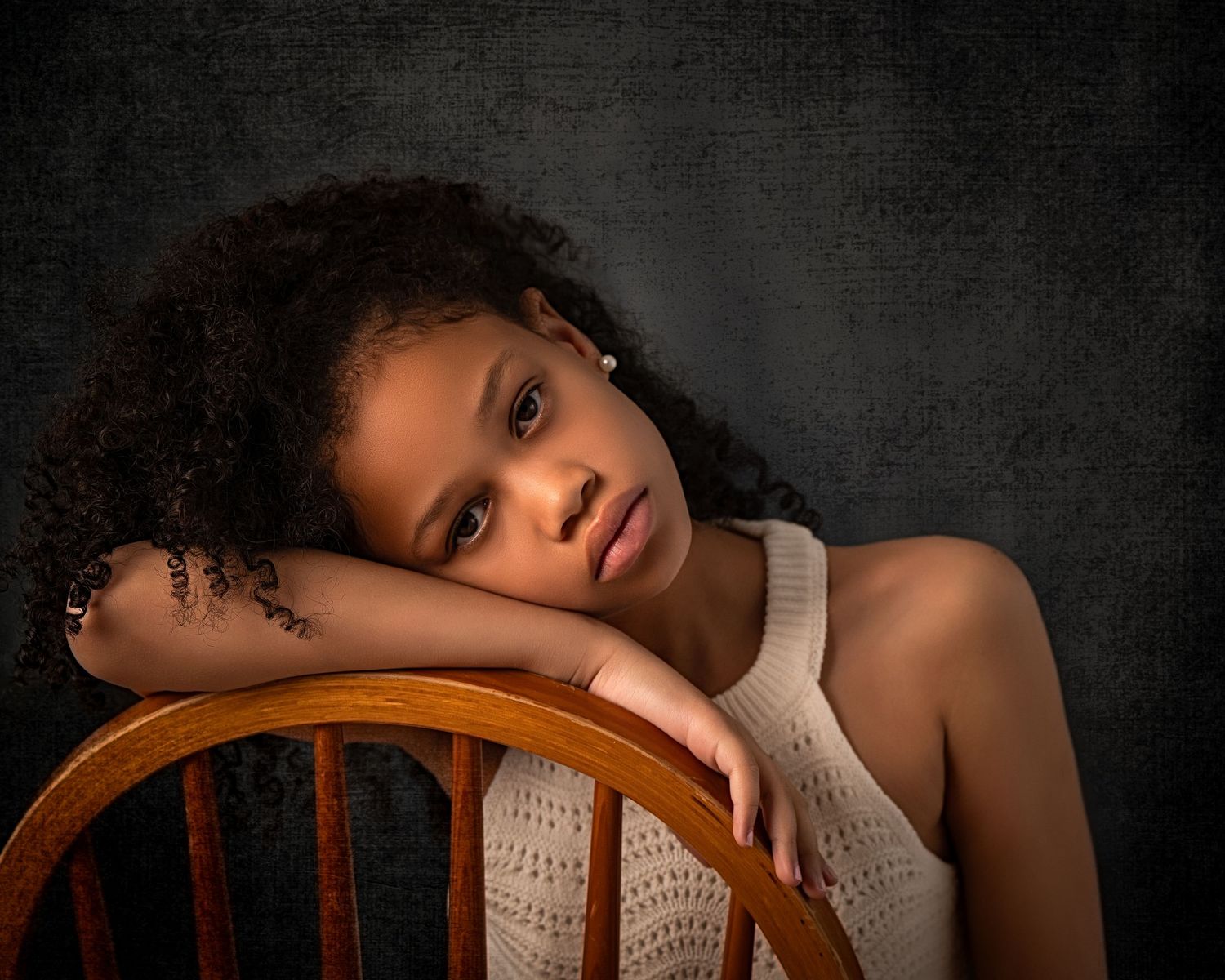Young girl resting her head on a wooden chair, softly lit against a dark studio background, wearing a light outfit with a thoughtful express