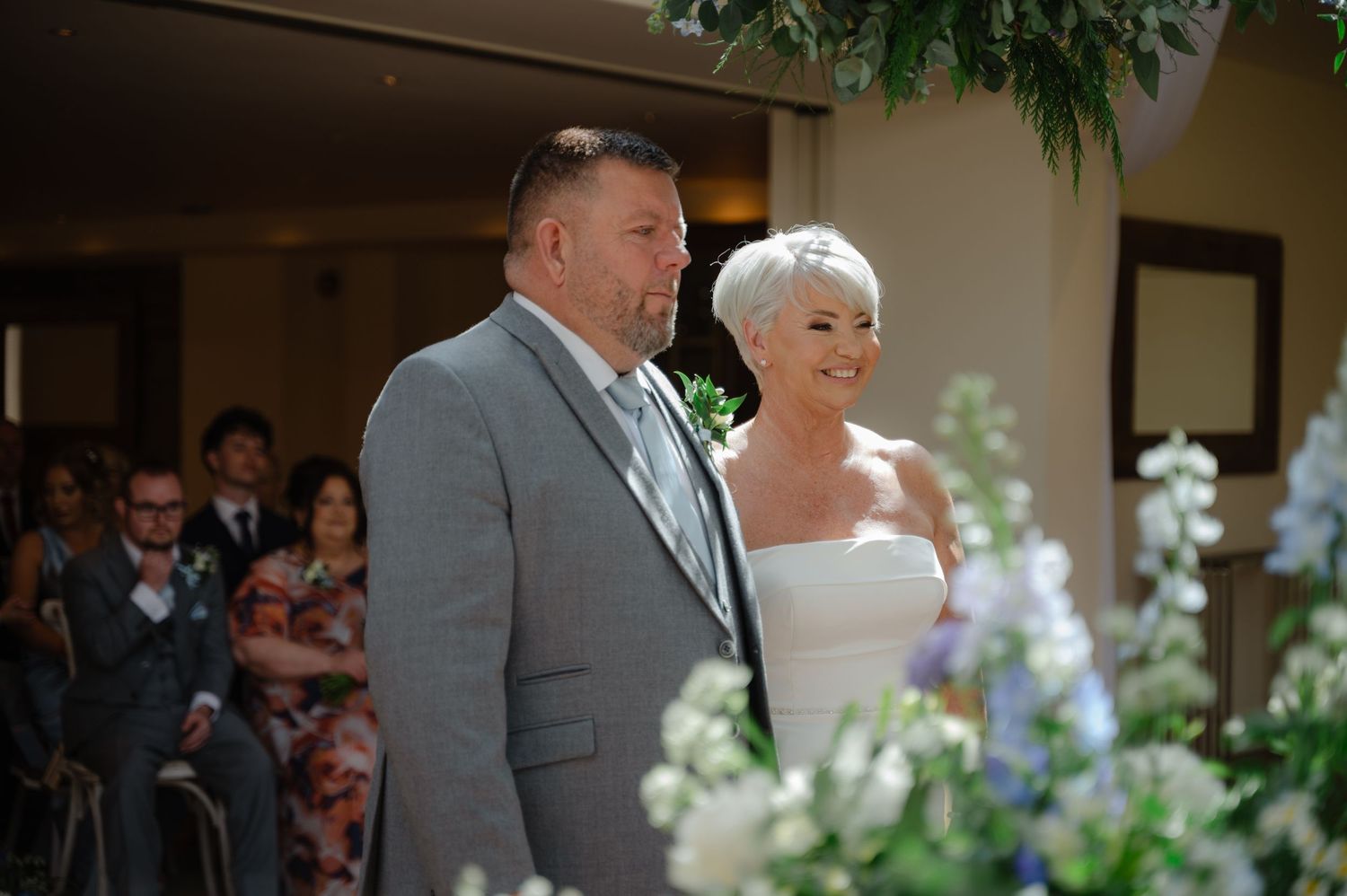 Bride and groom standing close and smiling at one another during the ceremony, framed by blue and white flowers at the front.