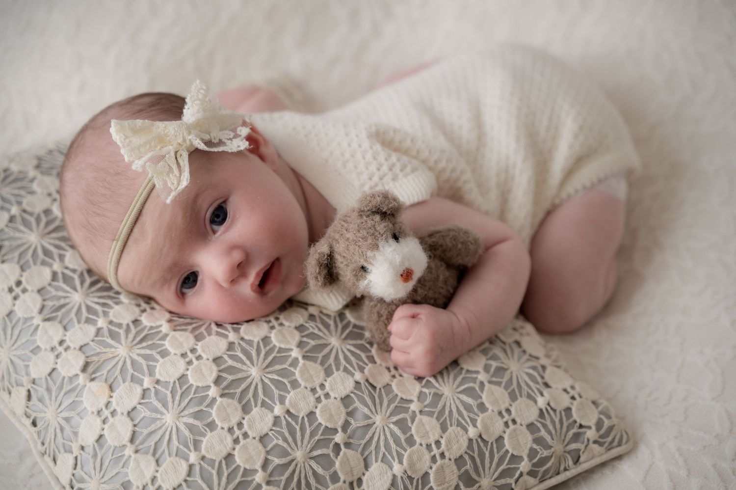 Newborn baby girl lying awake on a patterned pillow, wearing a bow headband and cuddling a small teddy bear, softly swaddled.