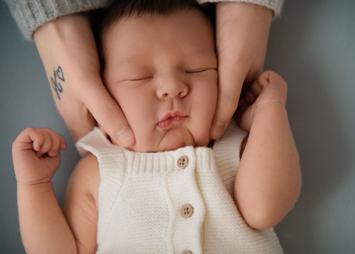 Newborn baby lying on a grey background, gently holding both parents’ fingers, wearing a sleeveless cream buttoned romper.