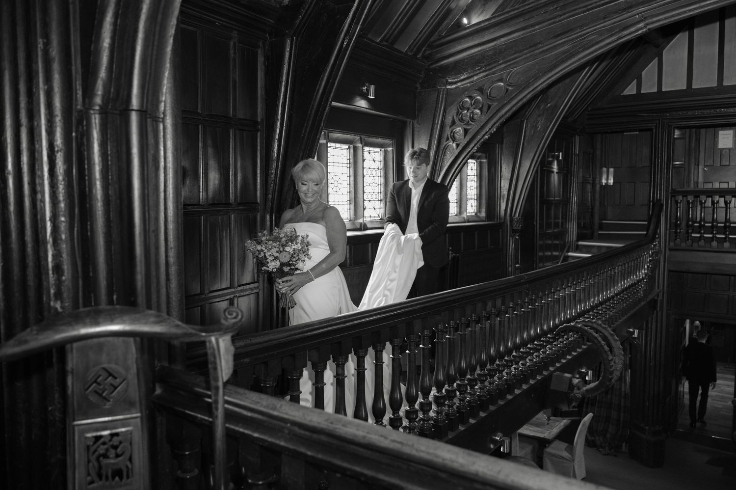 Dramatic black and white image of the bride standing on a grand wooden staircase, looking out over the balcony with her train trailing behin