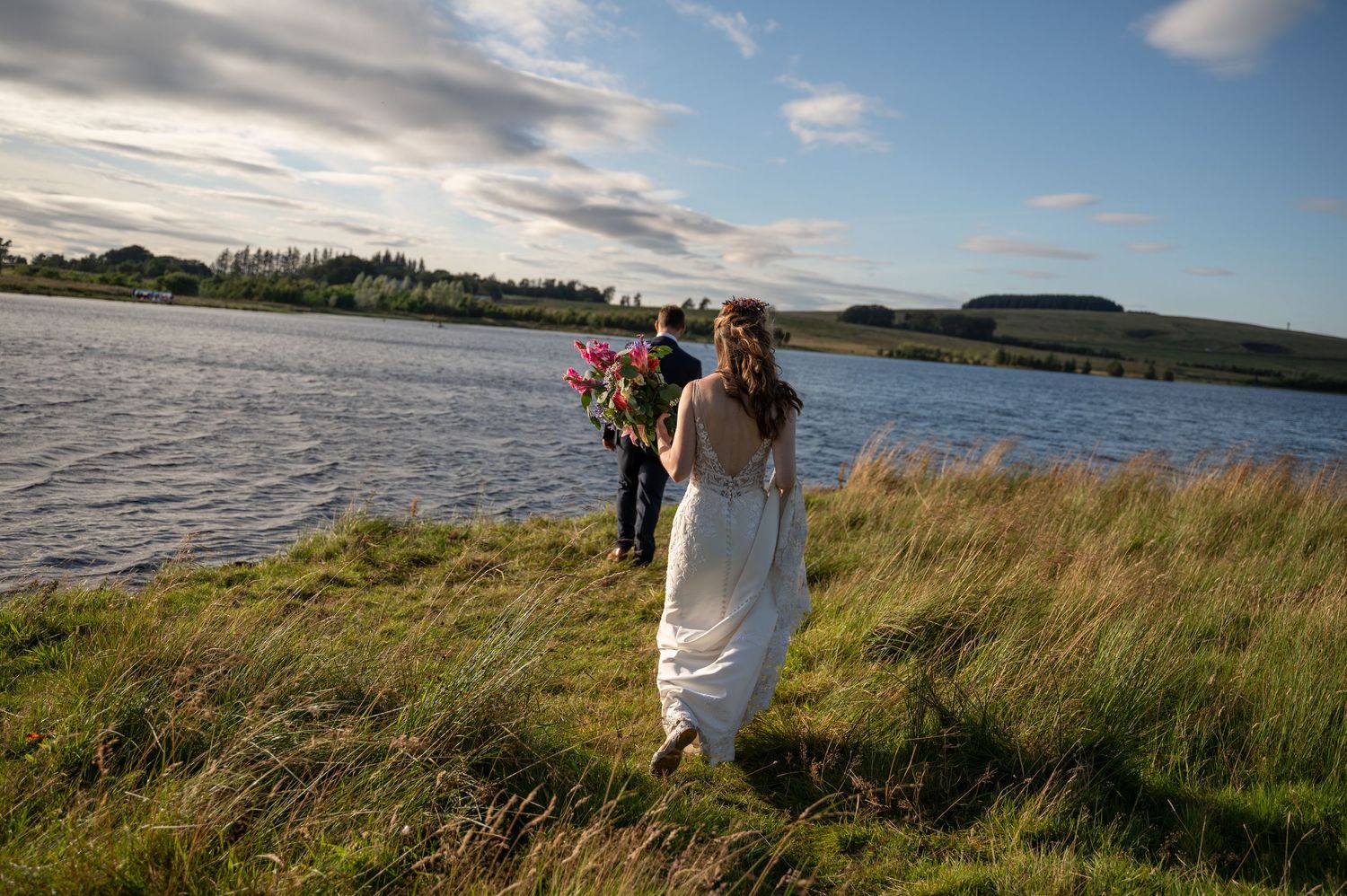 Bride walking through grass with bouquet near lakeside, wind blowing her dress