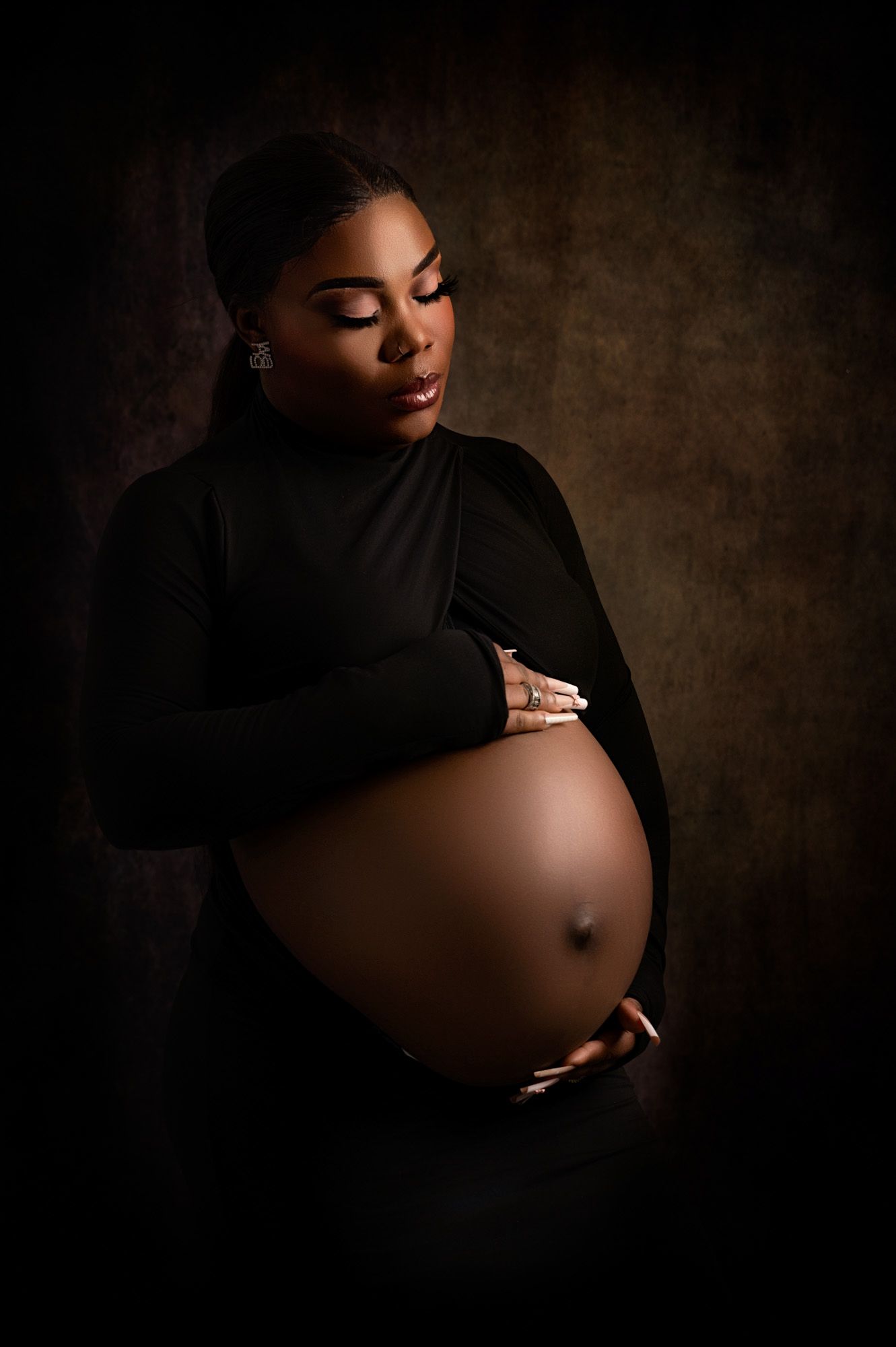 Pregnant woman in a dramatic dark outfit with a sheer panel revealing her belly, softly lit against a black background, exuding strength
