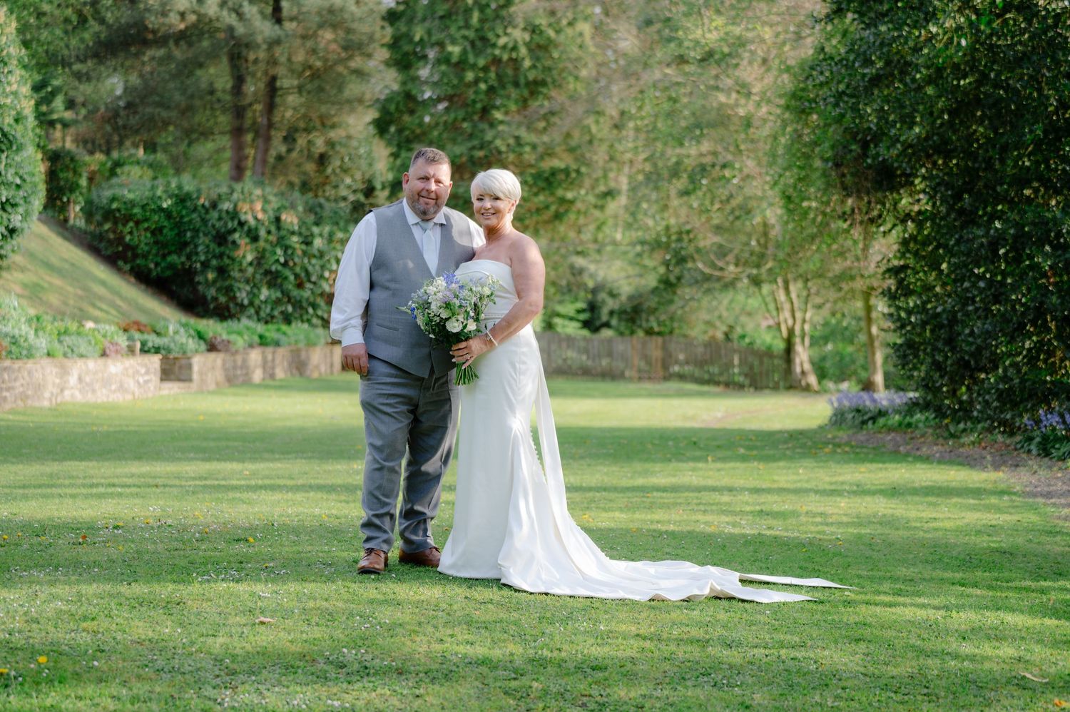 Bride and groom standing together on a green lawn, smiling at the camera, with the bride holding a bouquet and train flowing behind.