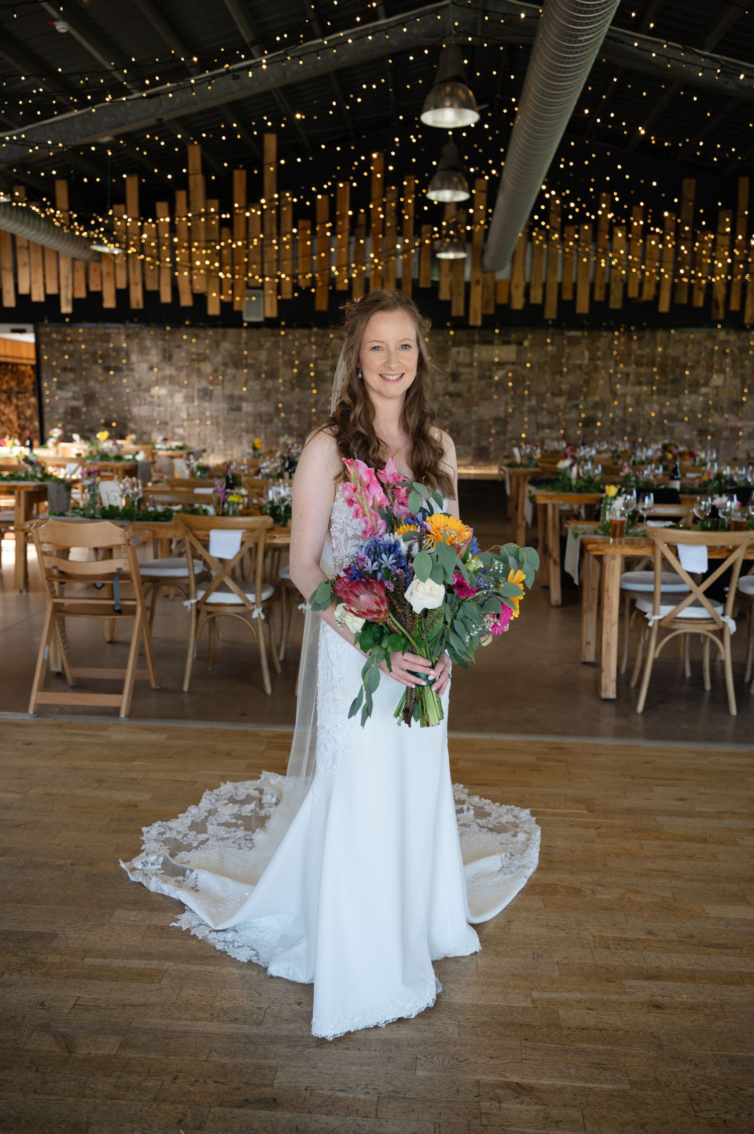 Bride holding bouquet while standing alone in a rustic barn wedding venue with hanging lights