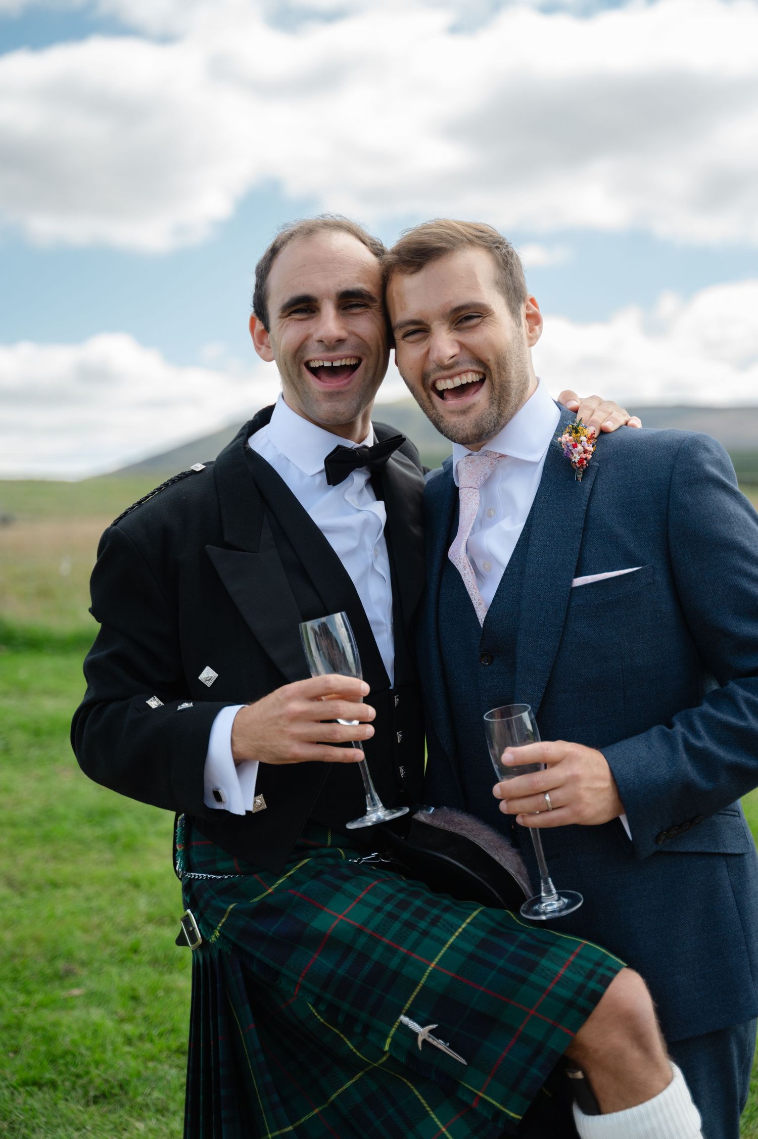 Groom and best man laughing and holding drinks in outdoor countryside setting