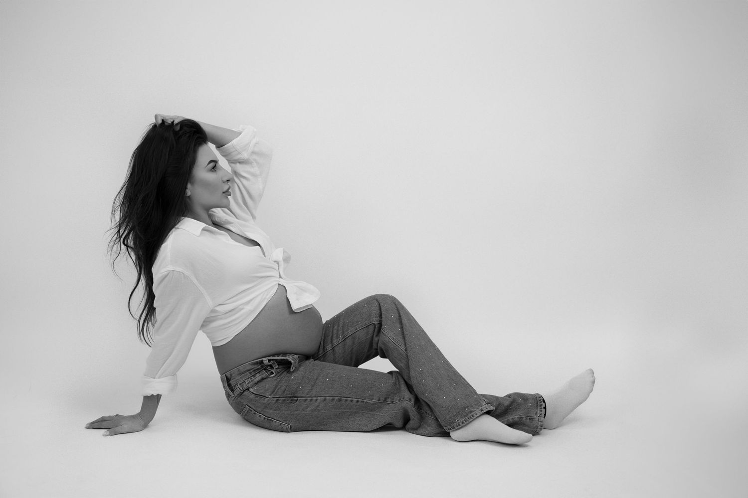 Pregnant woman in white top and jeans sits on floor in a minimalist white studio.