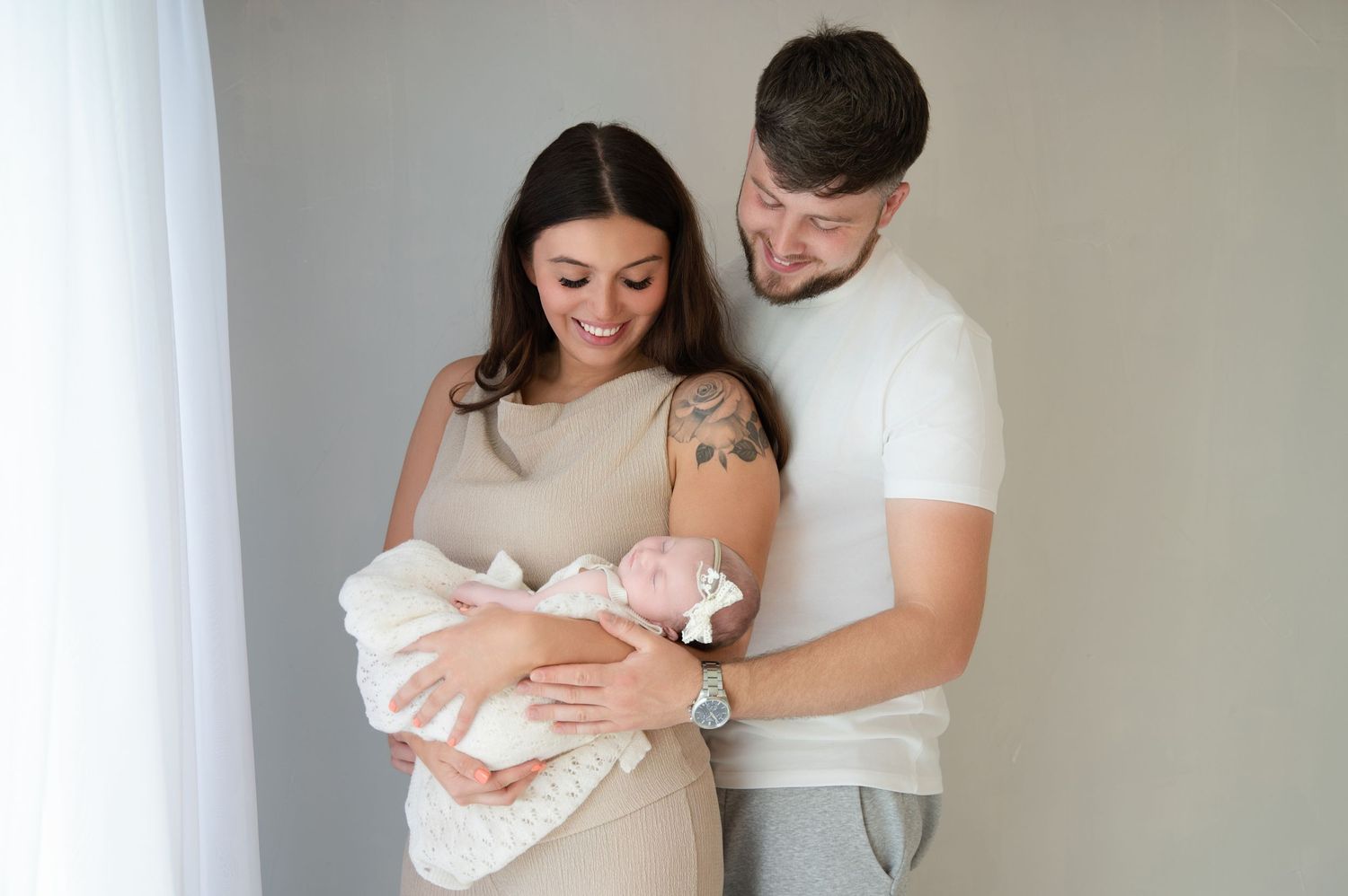 Young couple smiling as they hold their newborn baby girl in a neutral-toned studio setting, full of warmth and connection.