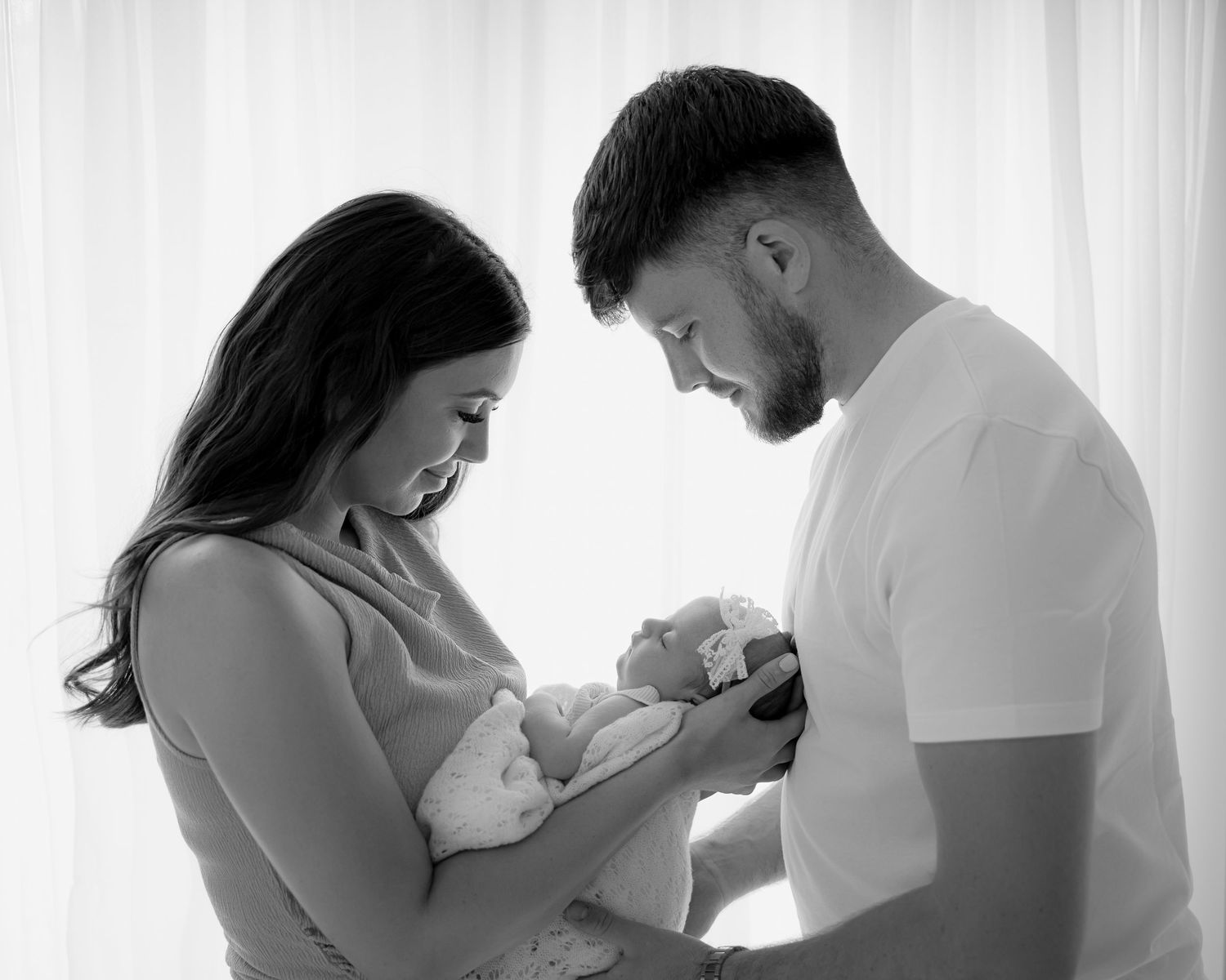 Young couple admiring their newborn baby, standing together in soft natural light, creating a warm and intimate moment.