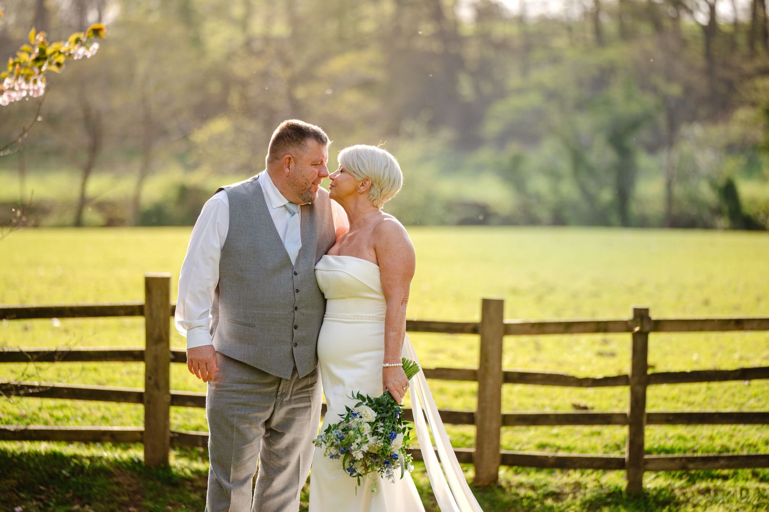 Bride and groom leaning in for a kiss in a sunlit field, standing in front of a wooden fence with soft greenery in the background.
