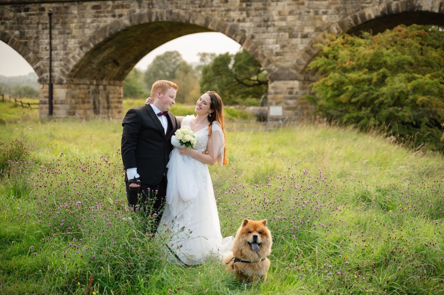 Bride and groom walking through meadow with their dog leading the way
