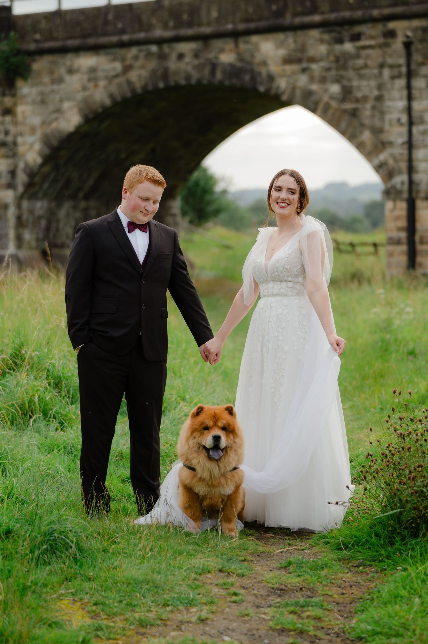 Bride and groom holding hands under a stone arch with their dog at their feet