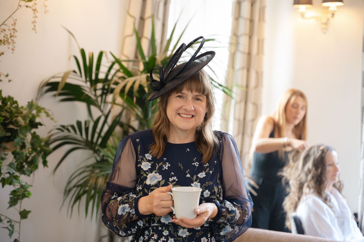 Mother of the bride smiling in a black and white outfit with a hat, holding a gift box during morning prep