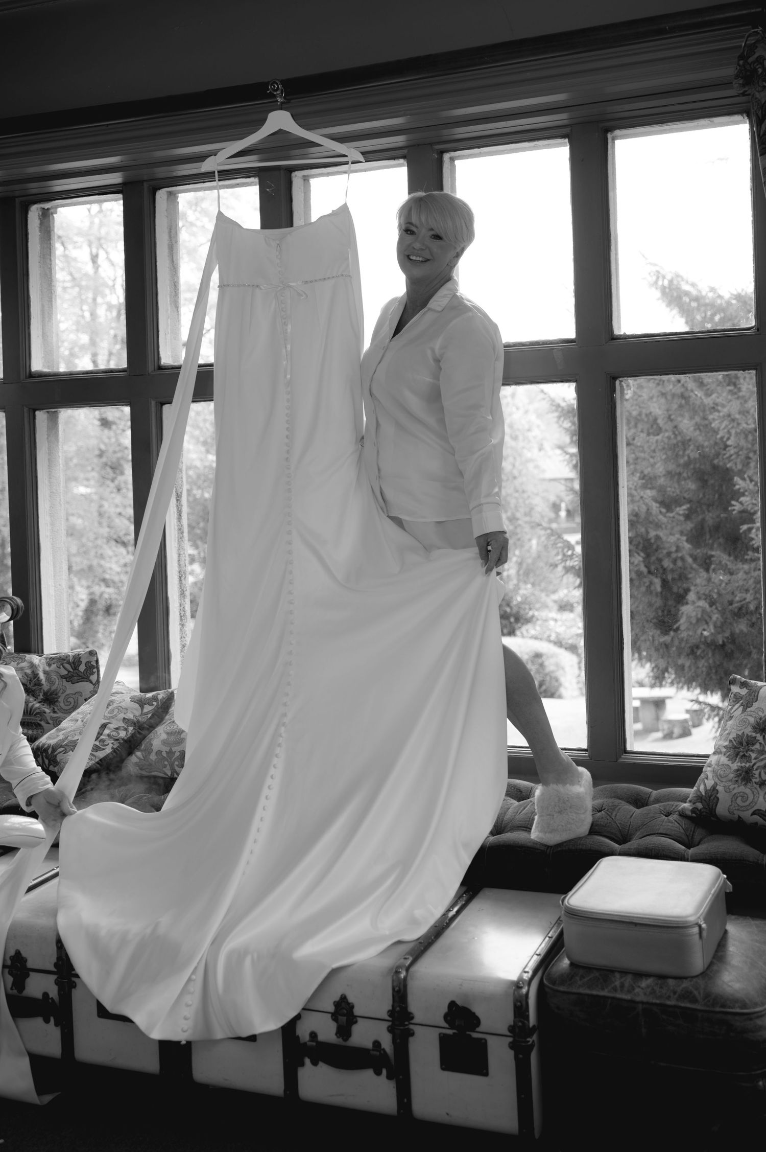 Black and white photo of a wedding dress hanging in front of a large window, capturing a quiet pre-ceremony moment in soft light.