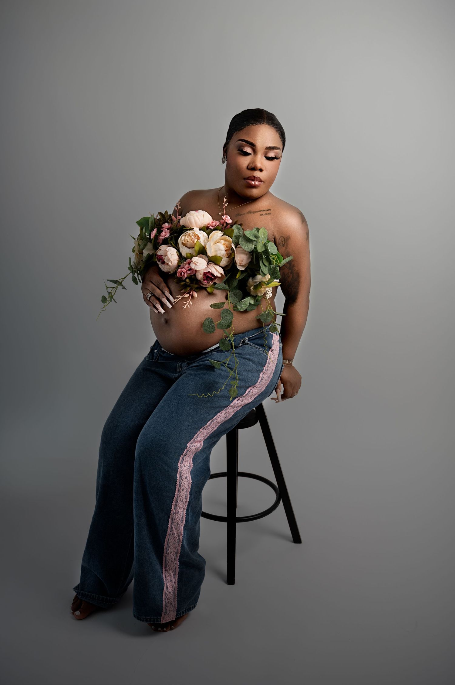 Seated pregnant woman in jeans and a floral top, holding a bouquet while sitting on a stool against a neutral backdrop.
