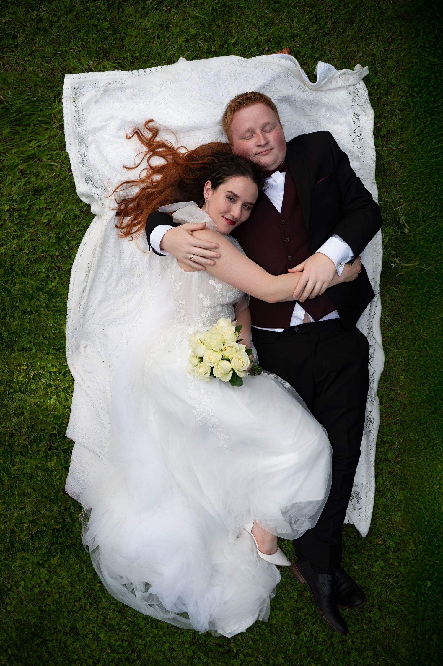 Bride and groom lying on the grass, holding each other and smiling up at the camera