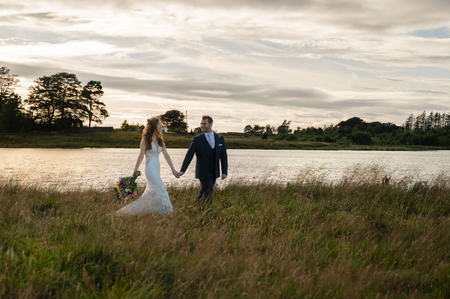 Bride and groom holding hands, walking through field at golden hour