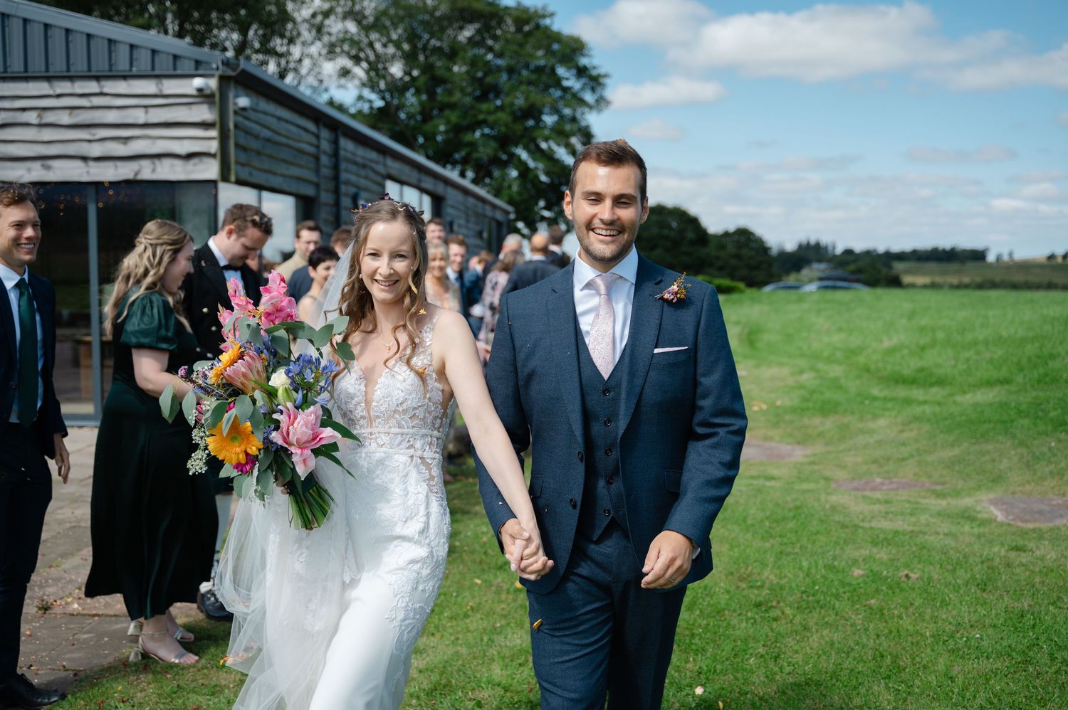 Bride and groom walking hand in hand outside, smiling with guests behind them