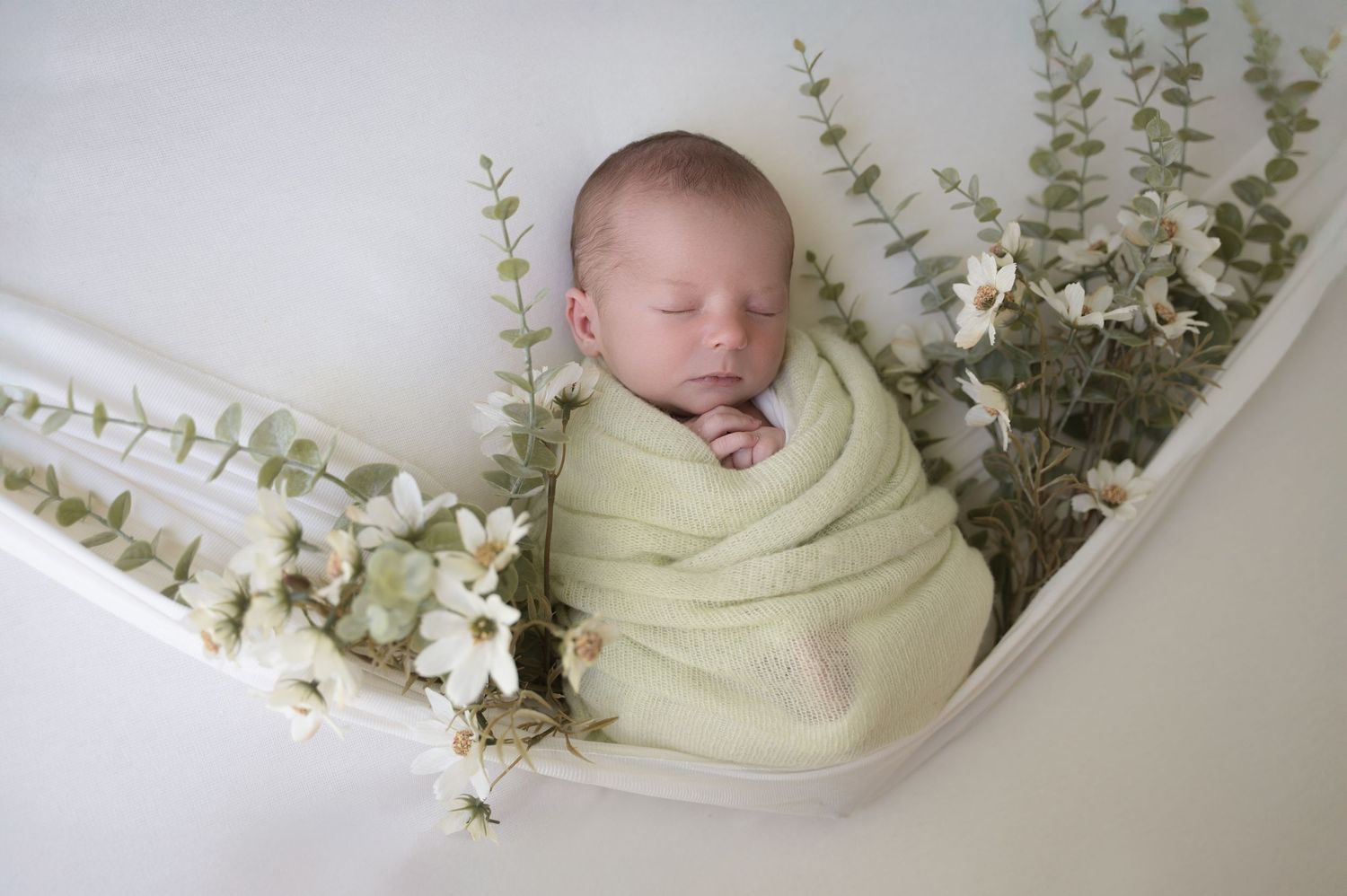 Newborn swaddled in a light green blanket, lying in a cream bowl with delicate white flowers arranged around.