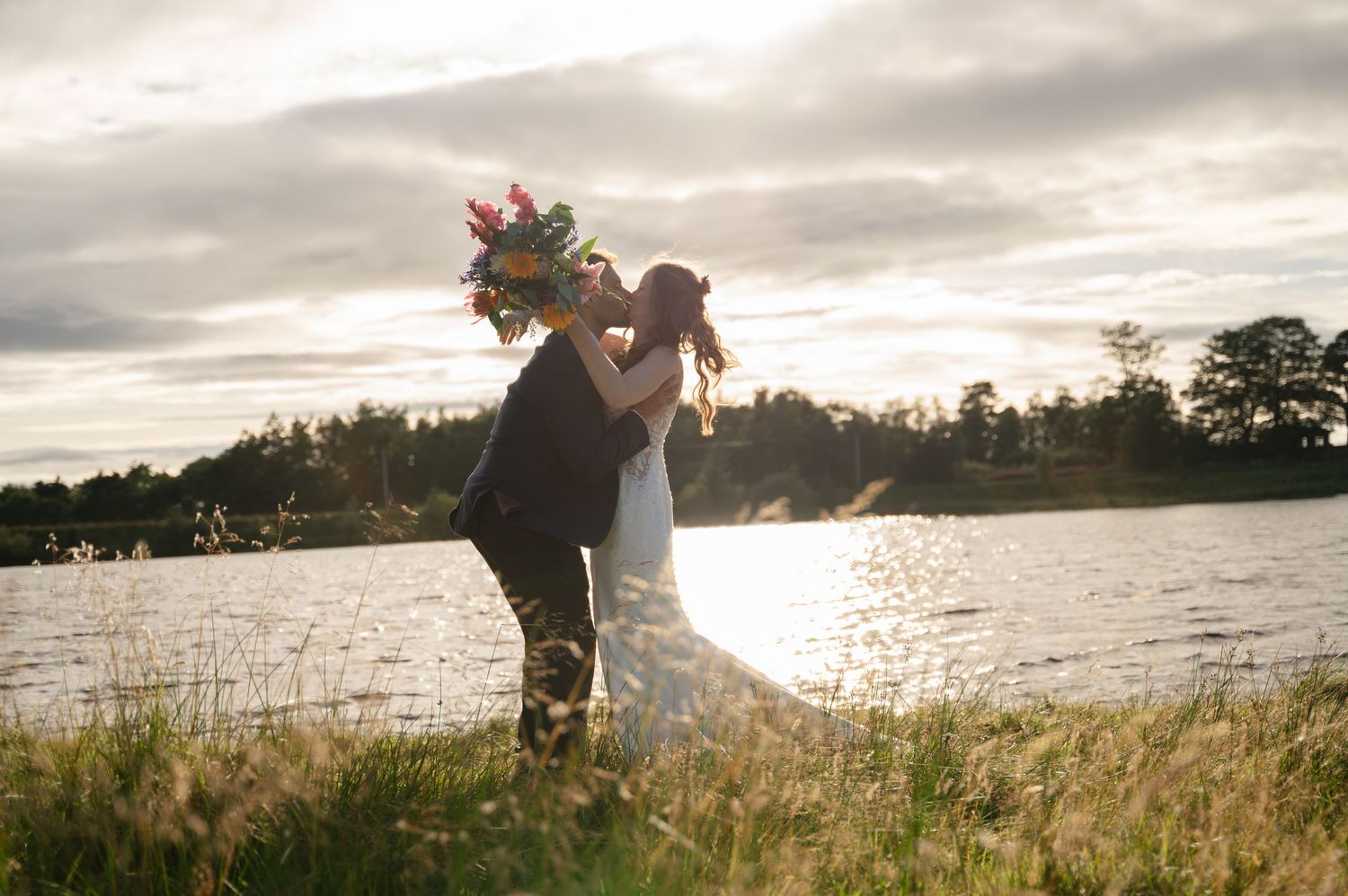 Bride and groom kissing beside a lake with golden sunset behind them